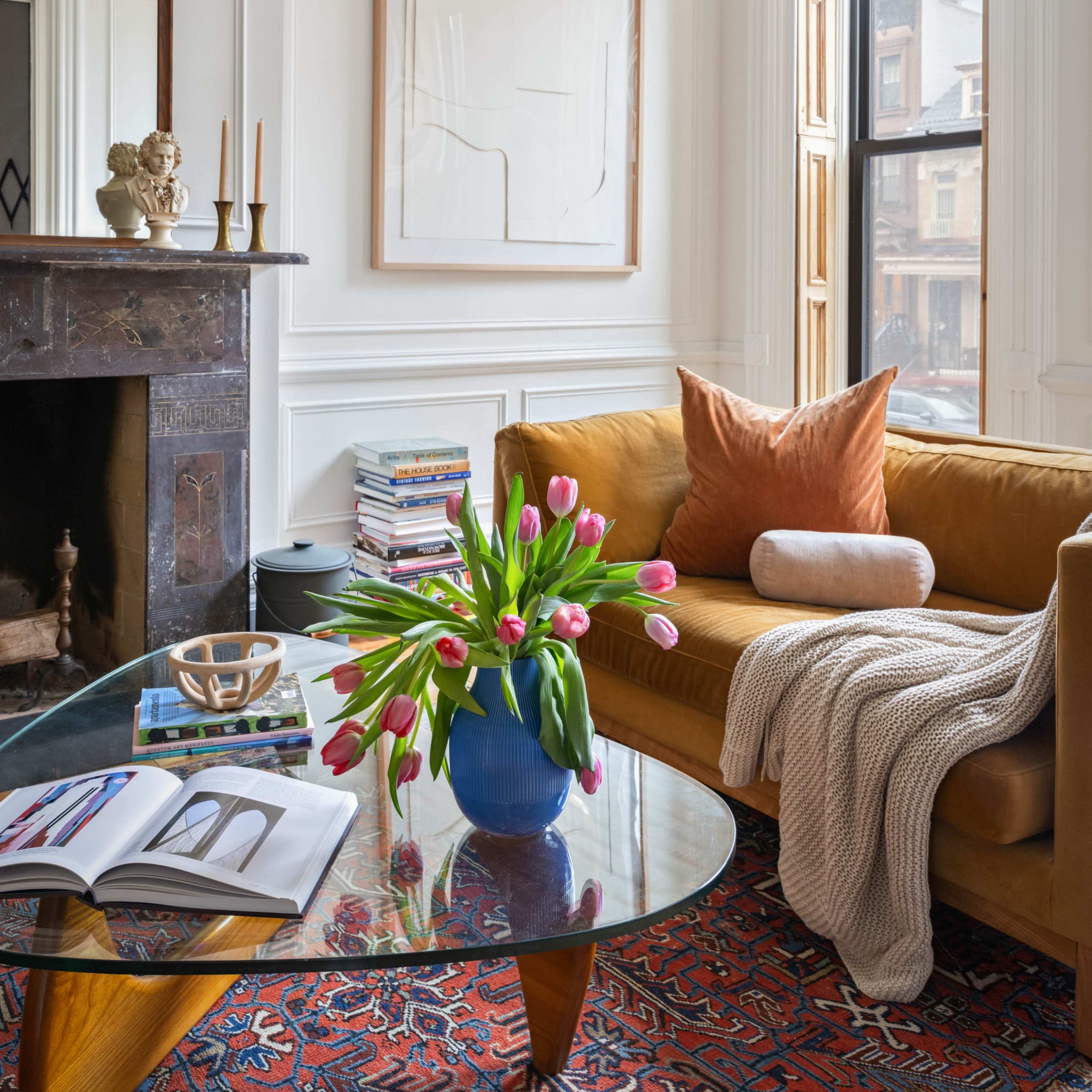 A cozy living room features a brown sofa, a glass coffee table with books and a magazine, a vase of pink tulips, and large windows allowing natural light to enter.