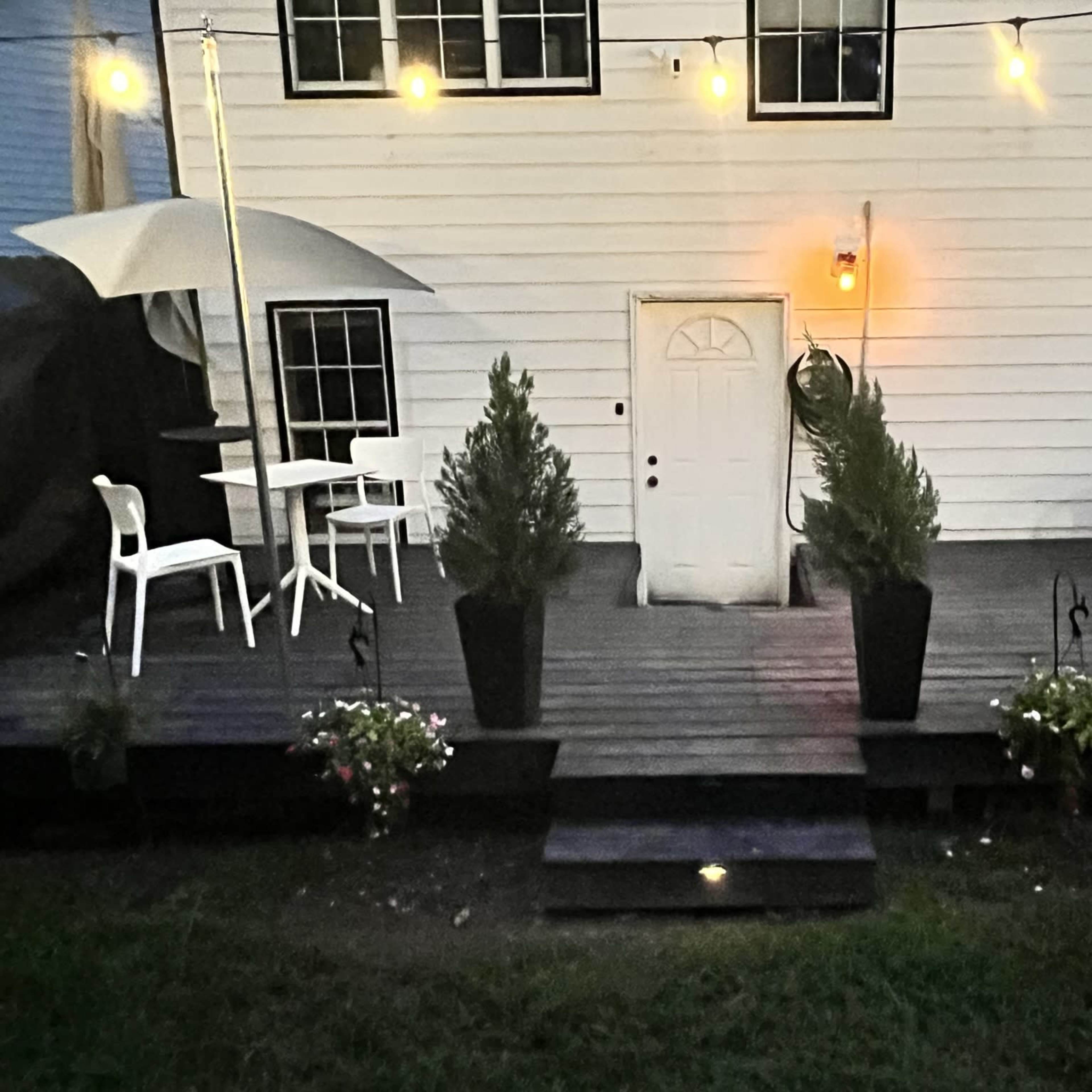 A wooden deck features a white door at the center, surrounded by potted plants, outdoor seating, and string lights above.