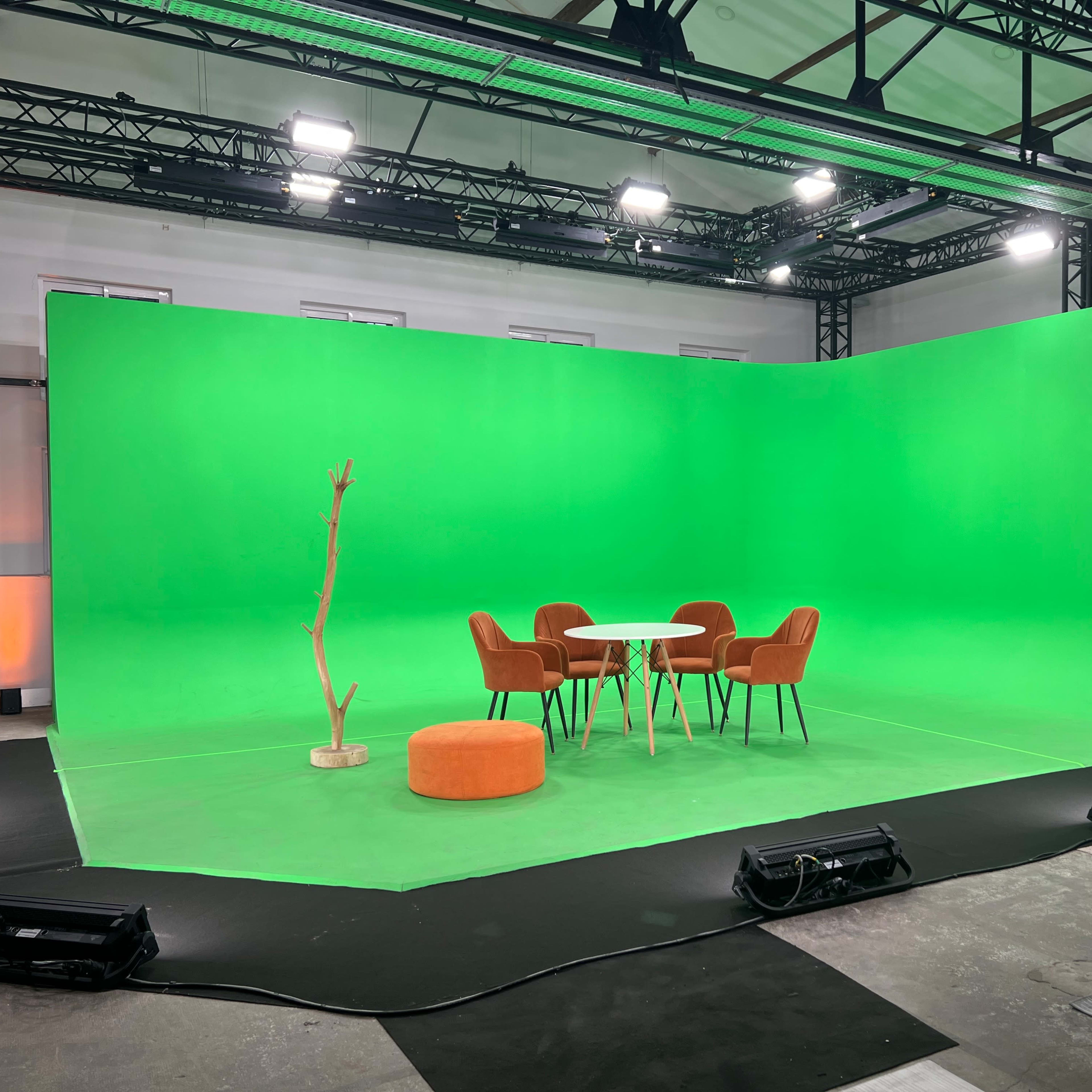 A green screen studio featuring a circular orange seating, a small white table, and four brown chairs arranged under overhead lighting.