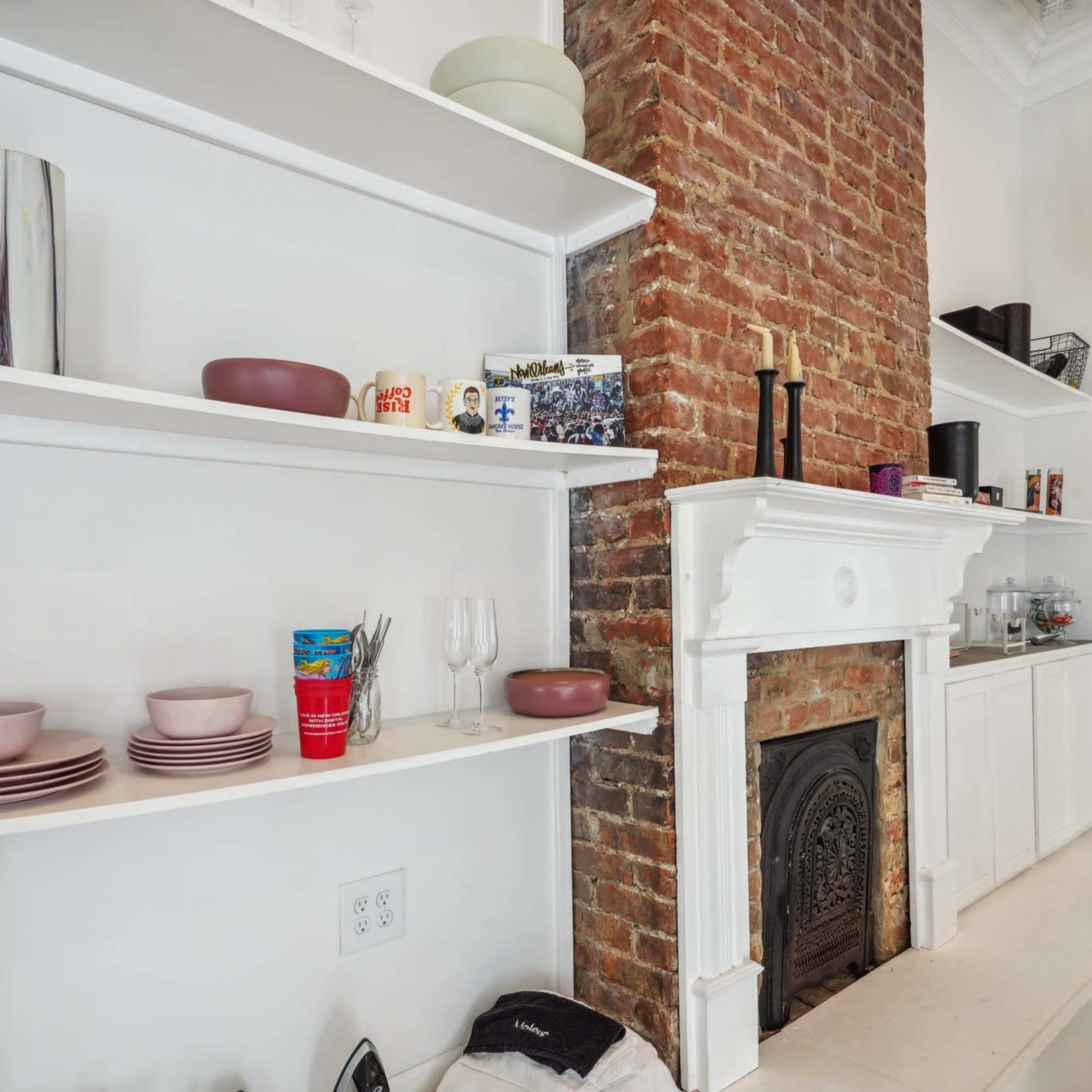 A white shelf holding dishware and glassware is positioned next to a brick fireplace in a well-lit room.