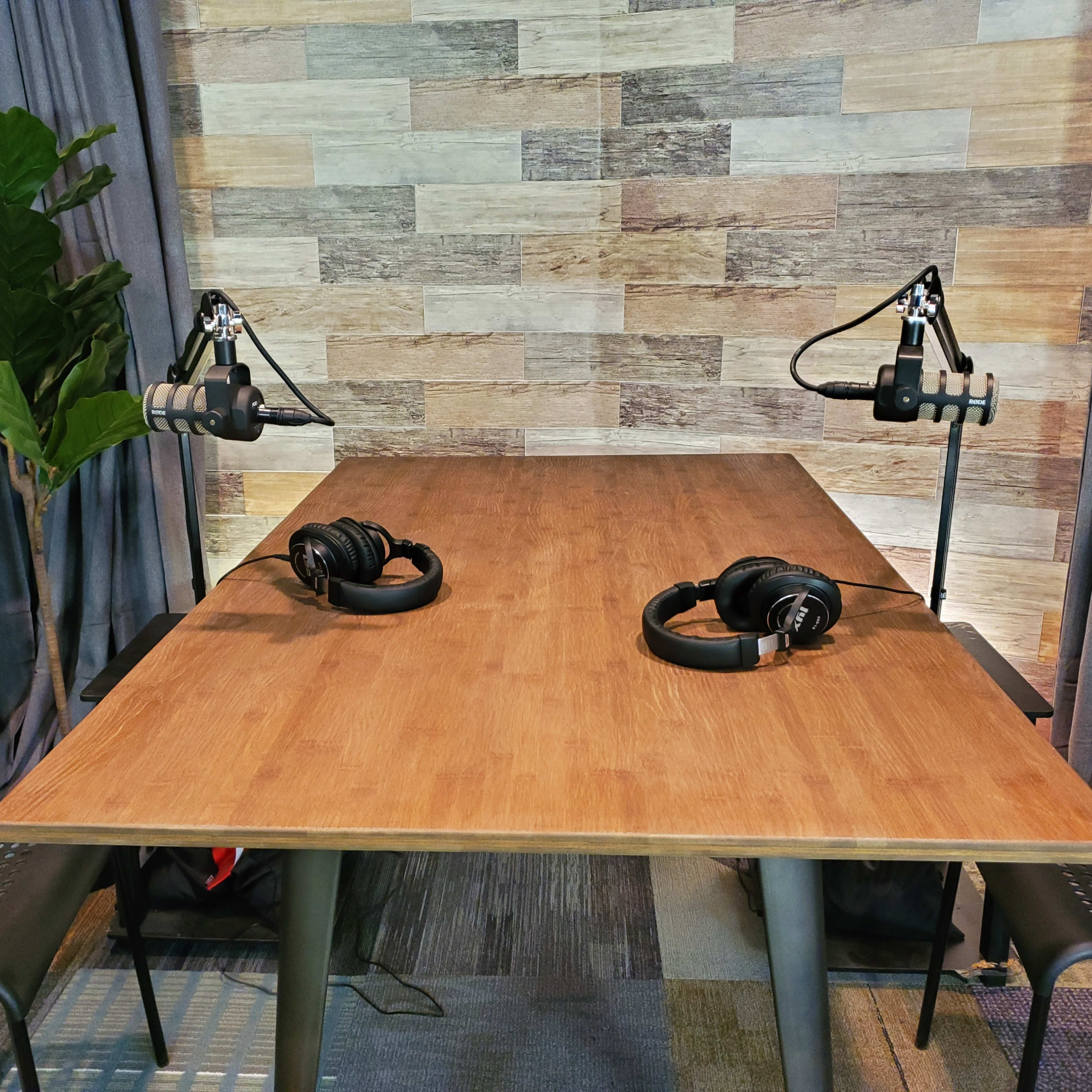 A wooden table with two microphones and headphones is set up in a room with a wooden panel background and potted plants on either side.