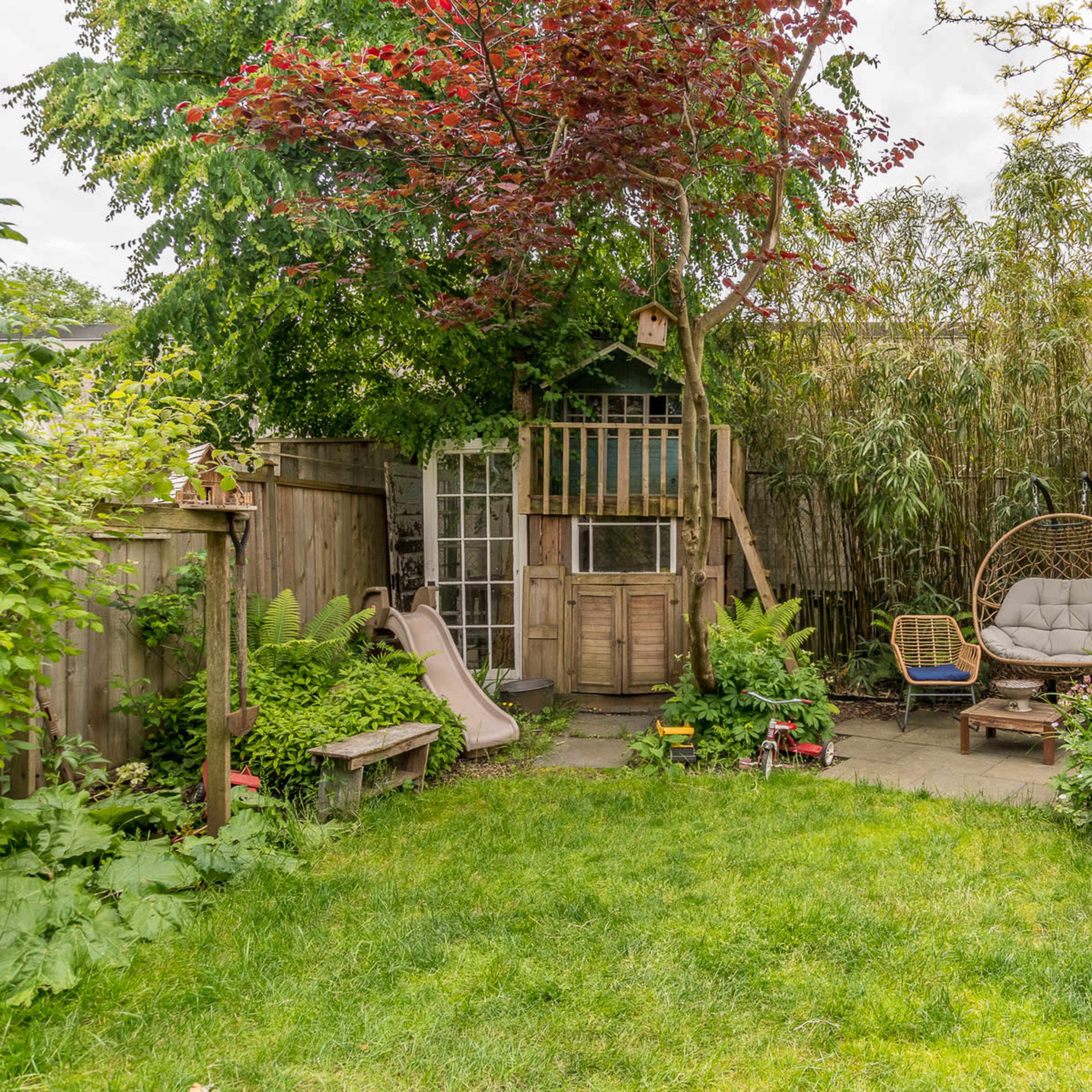 A backyard scene features a playground structure, a slide, a hanging chair, and lush greenery surrounding a stone patio.