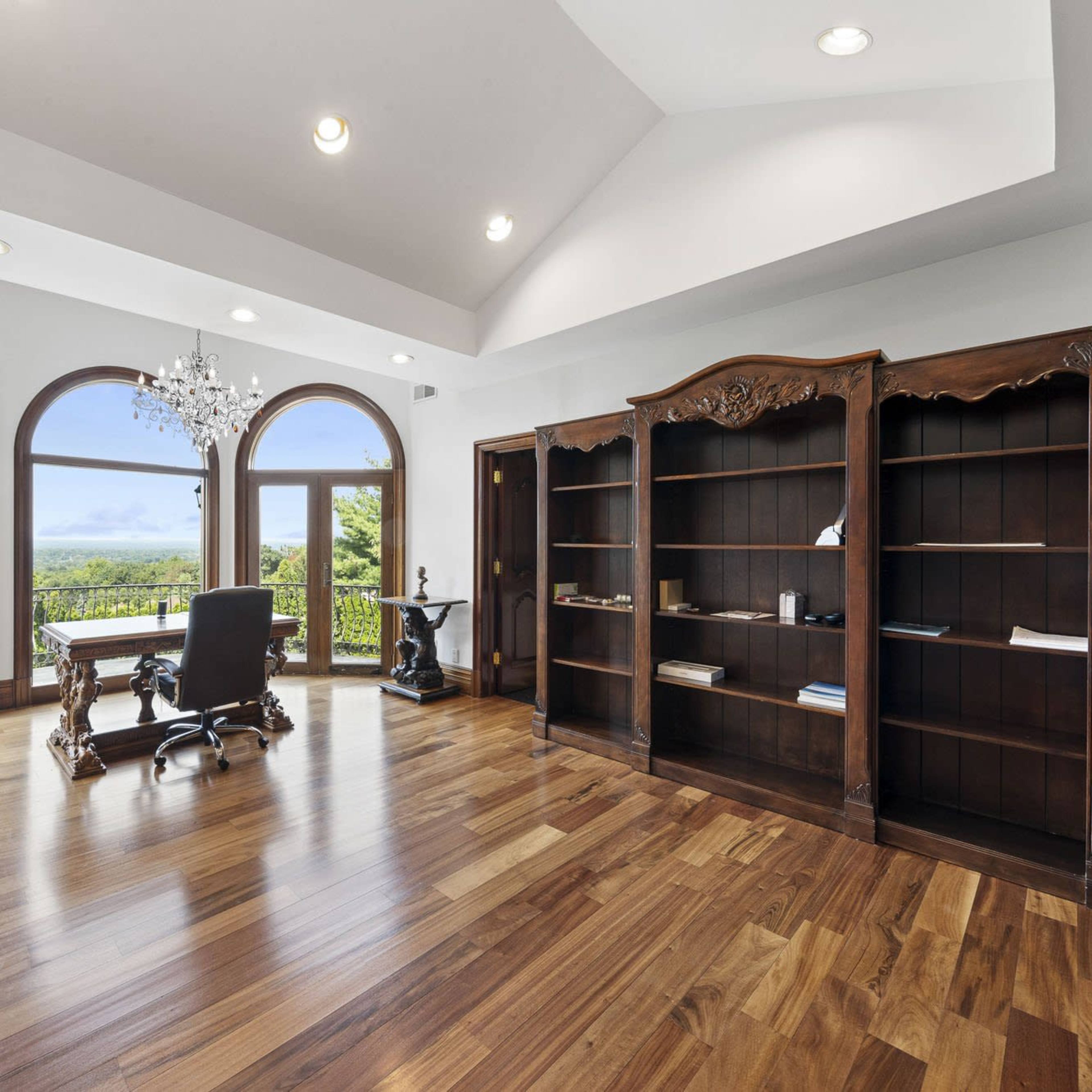 A wood-floored office space with large arched windows, a chandelier, a desk, and a bookshelf.
