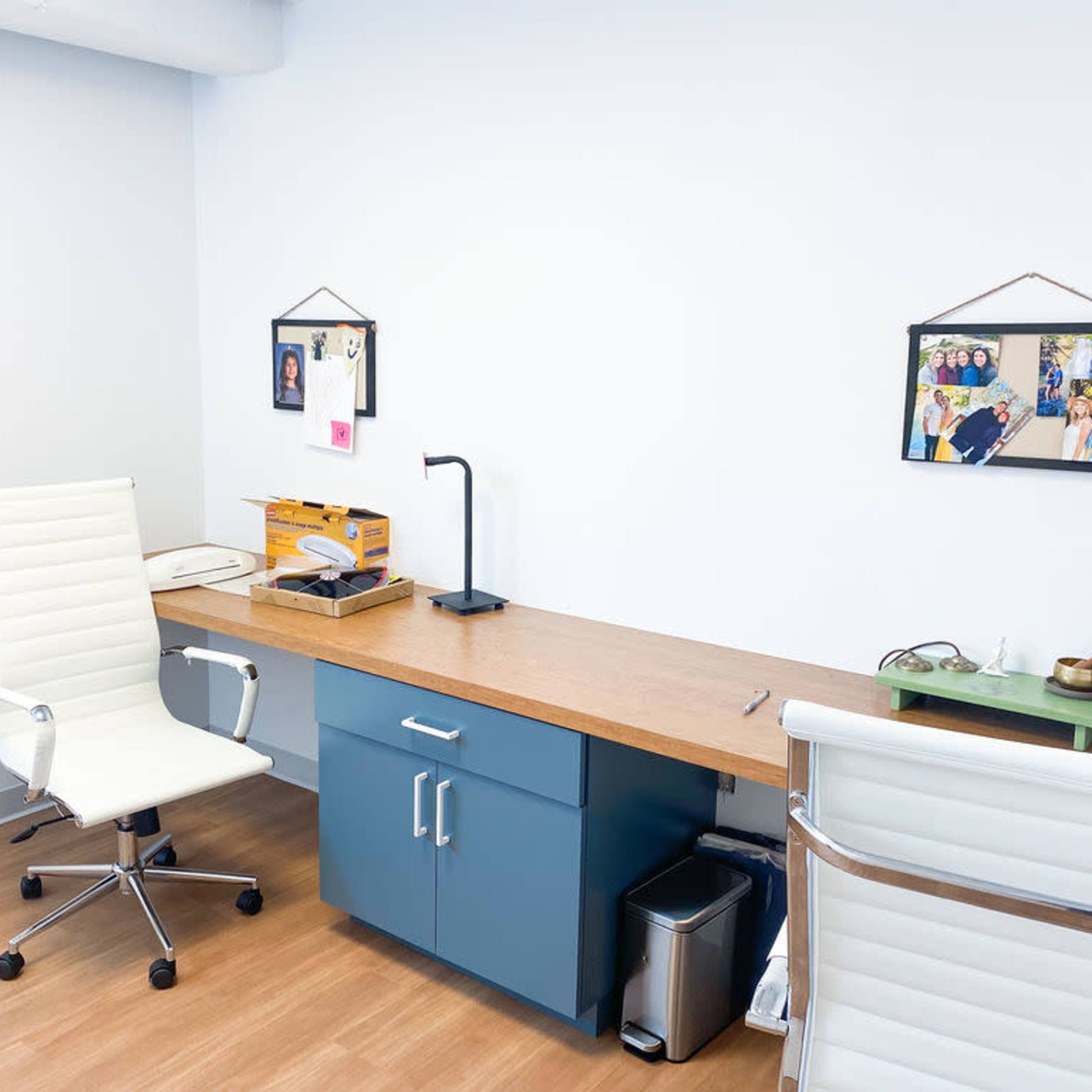 A minimalist office space with a wooden desk, two white chairs, a small trash bin, and framed photos on the walls.