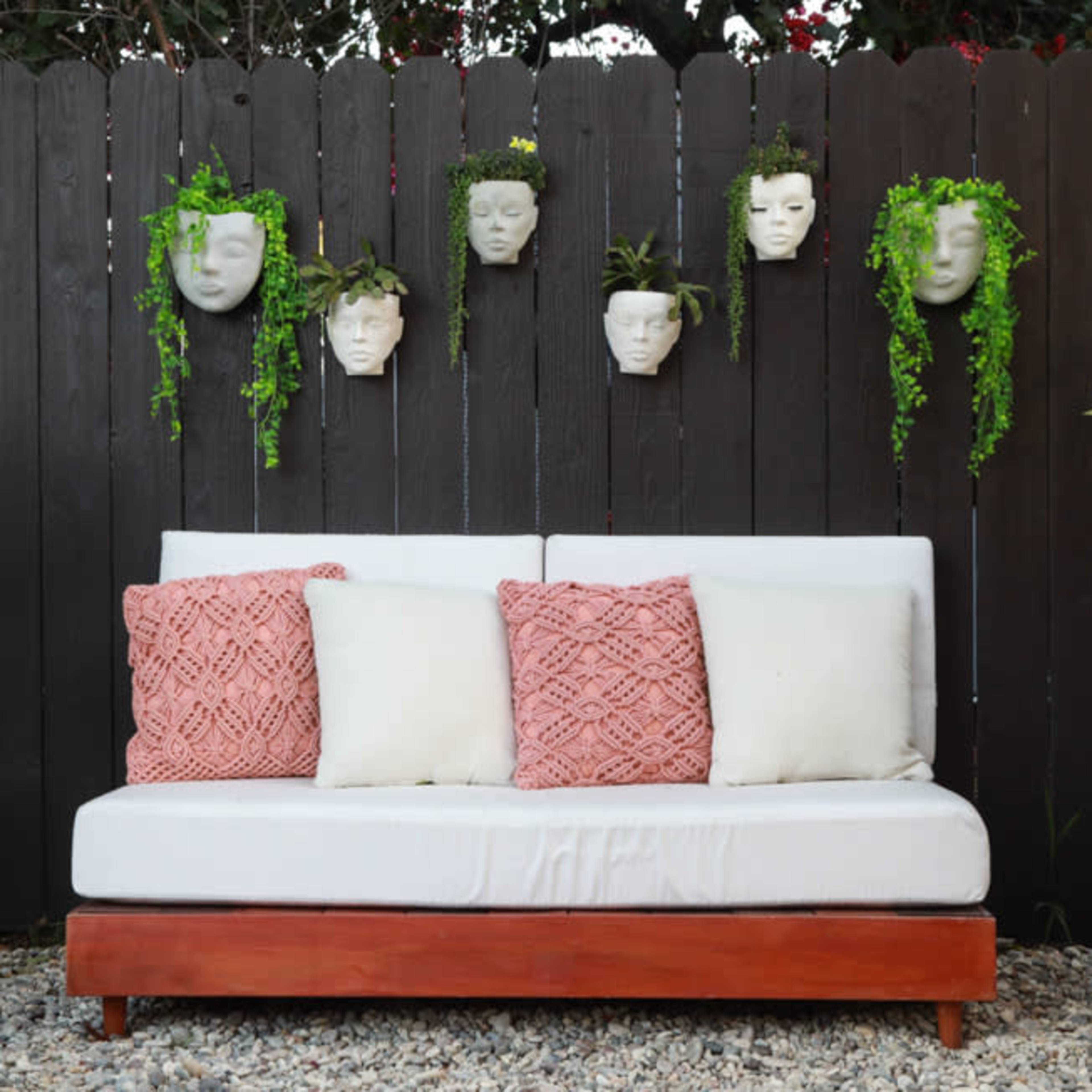 A white couch with cushions is positioned in front of a dark fence adorned with five planters shaped like faces, each featuring trailing green plants.