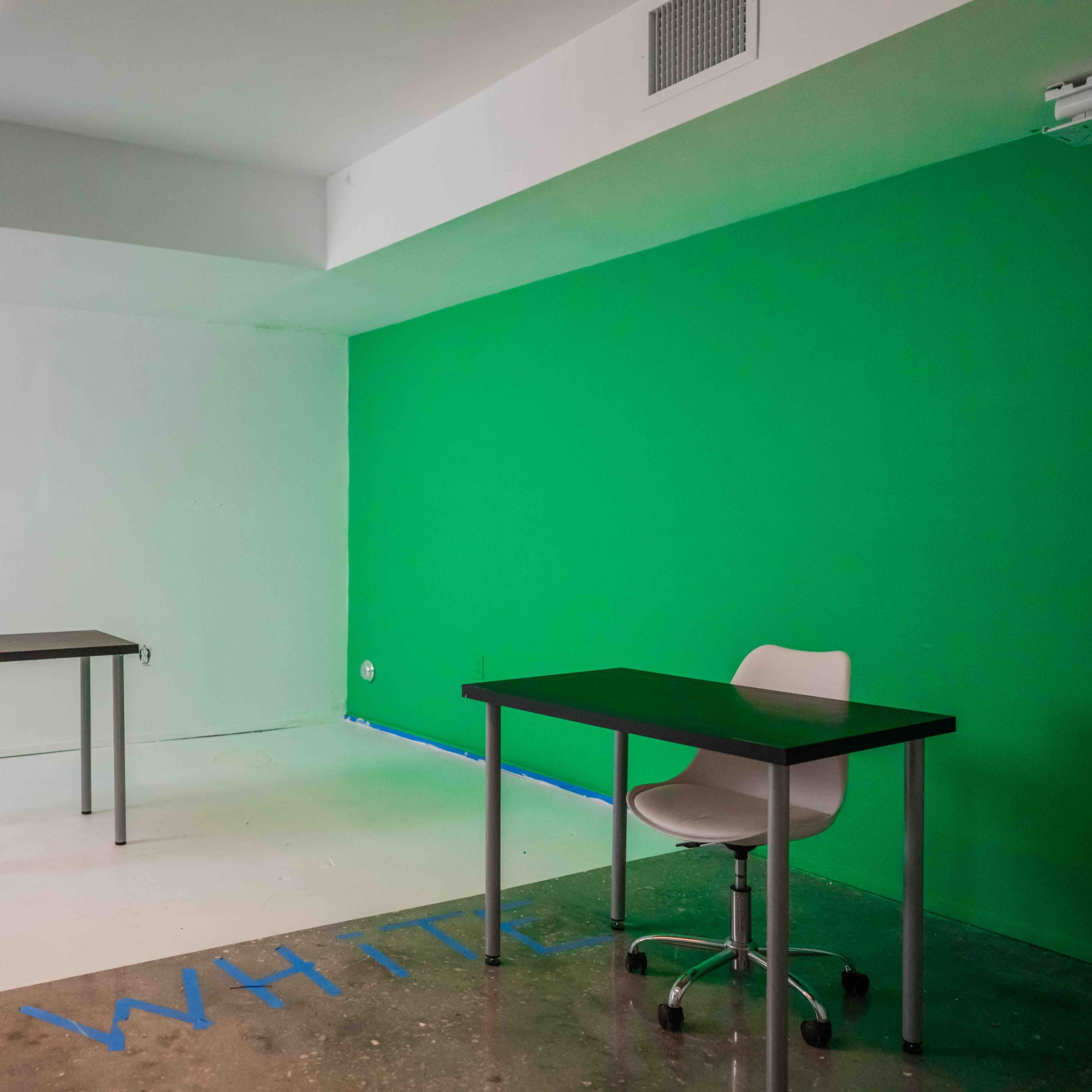 A room with a green wall, two tables, and a chair, with one table positioned in the center and the other against the wall.