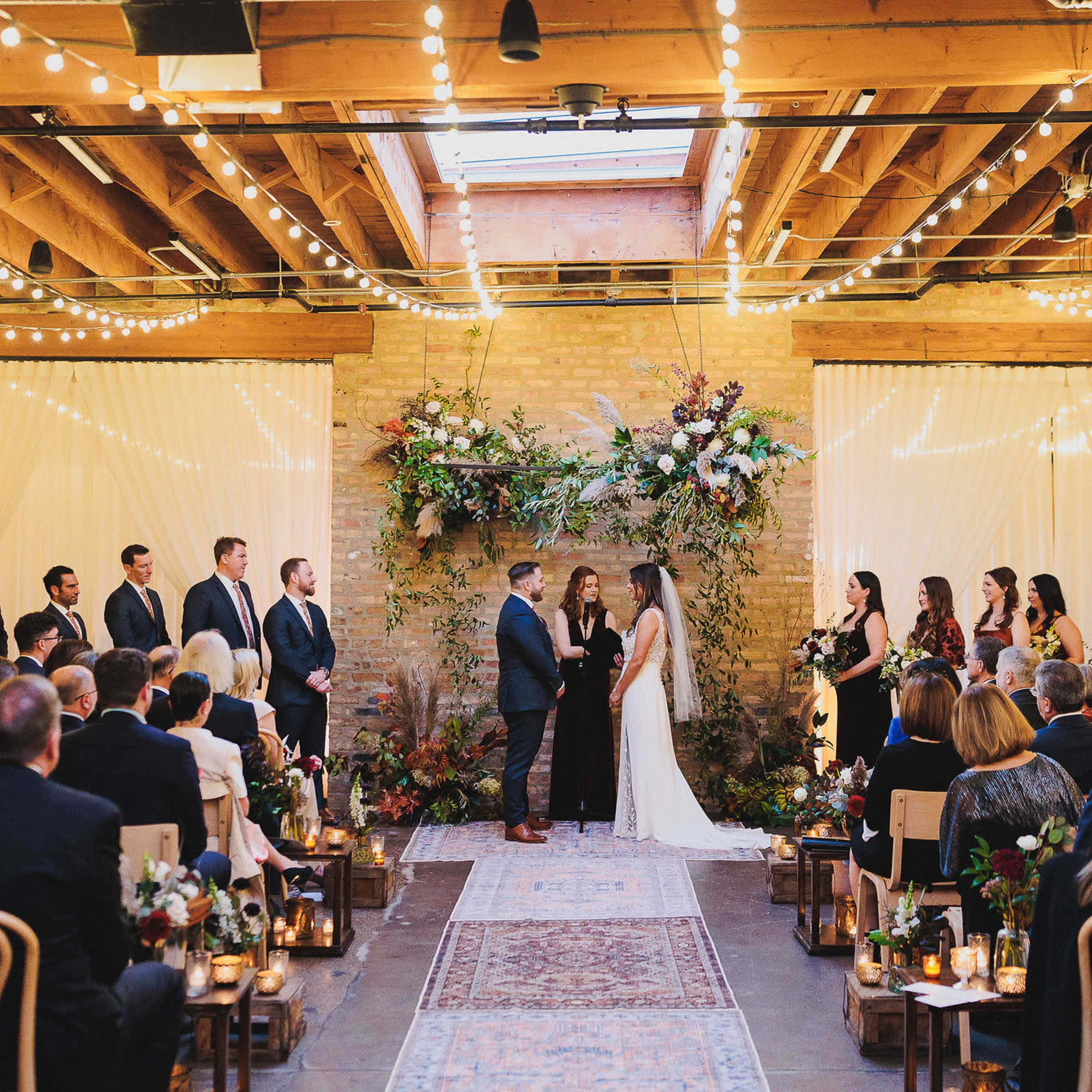 A couple stands in front of a floral arch during their wedding ceremony, surrounded by guests seated in rows.