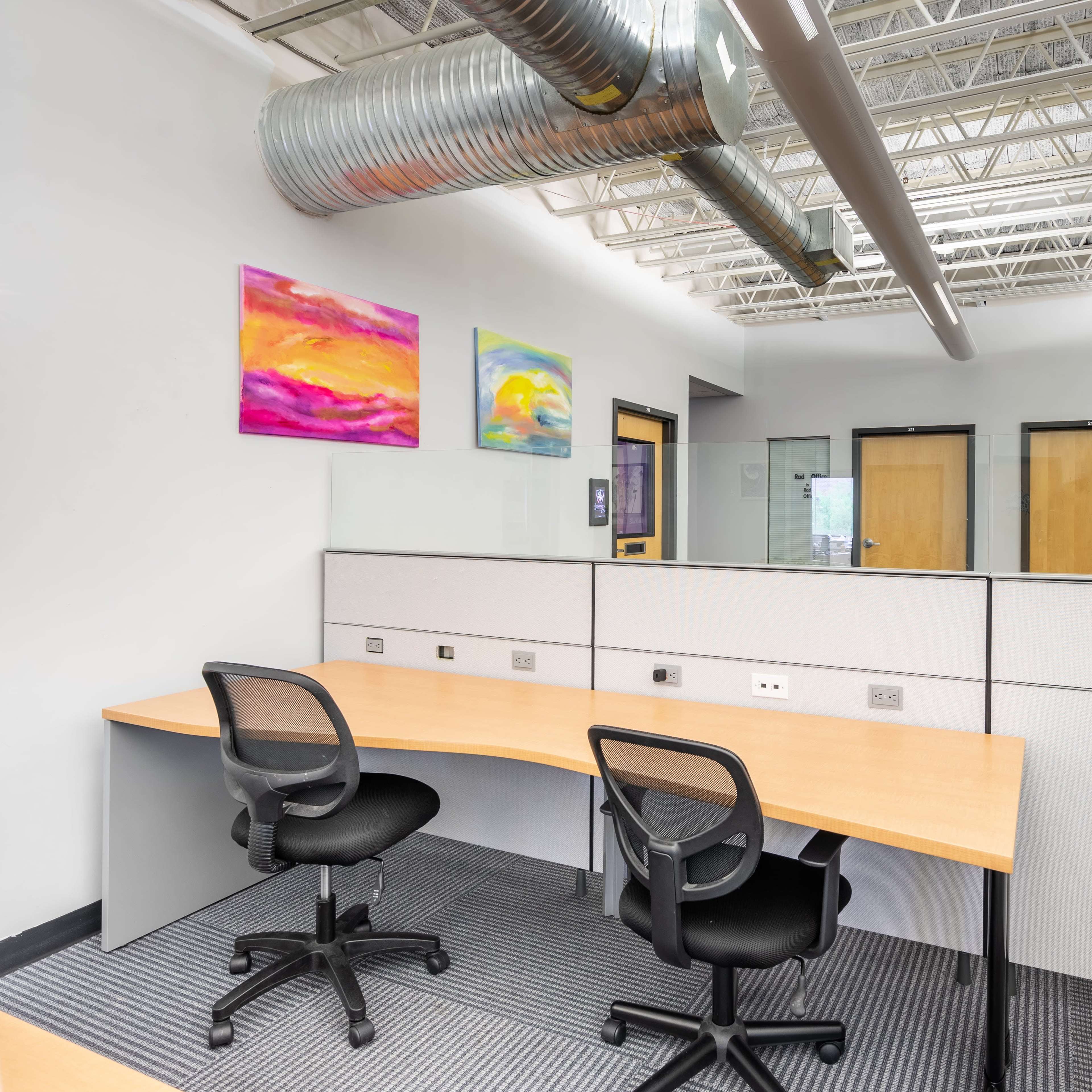 An office space with two empty desks and glass partitions, featuring colorful artwork on the walls.