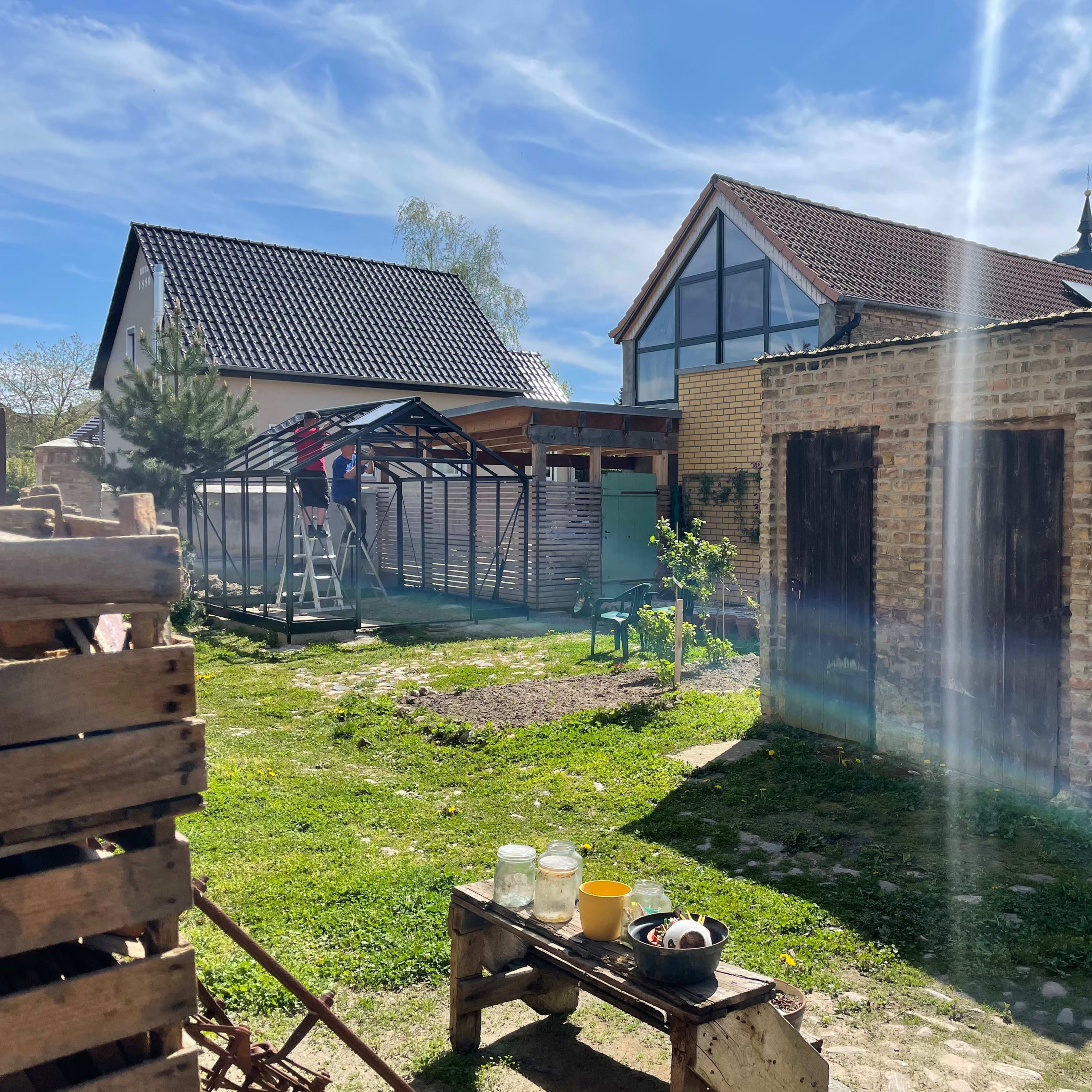 A backyard scene features a small stone shed, a wooden structure under construction, and a nearby house, all set against a clear blue sky.