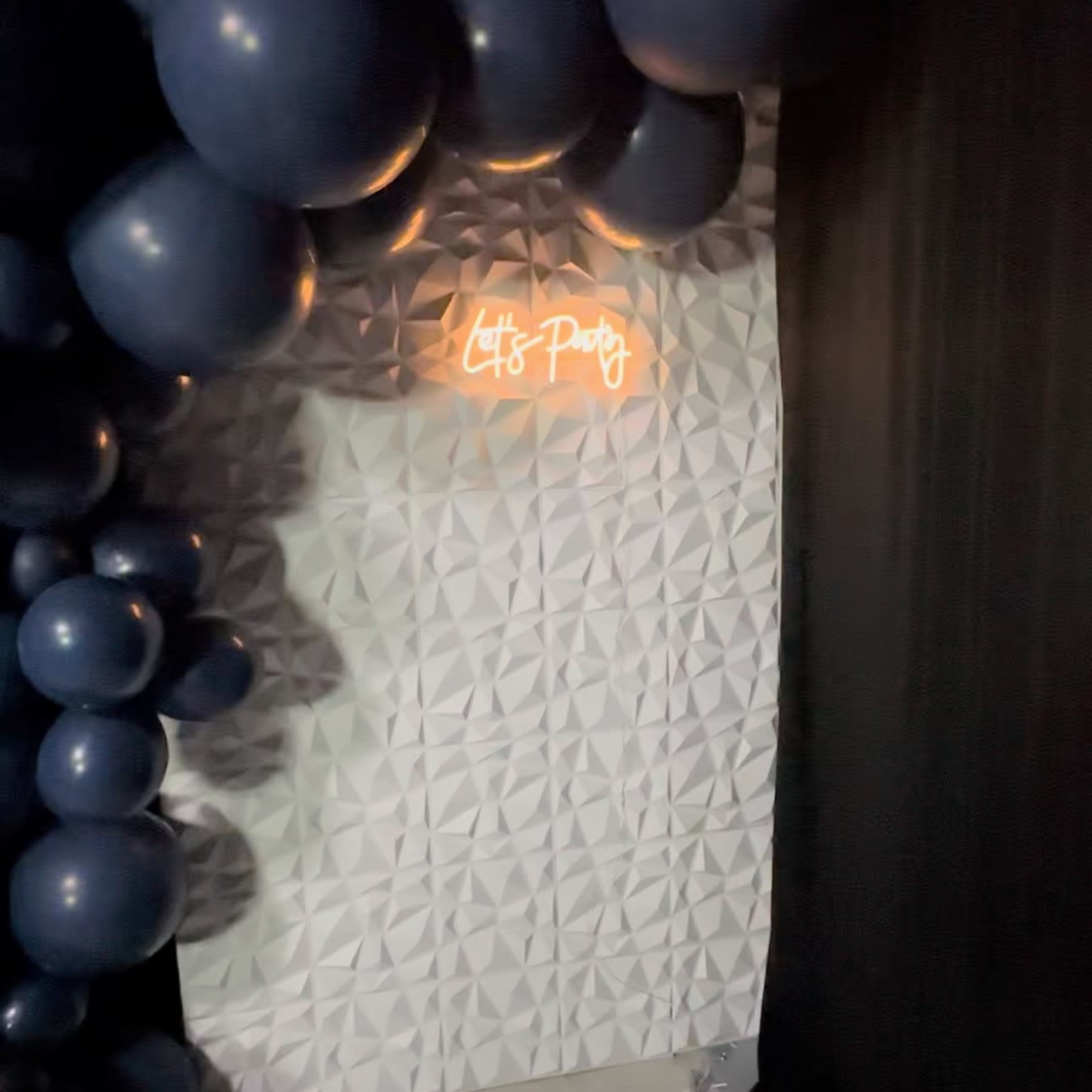 A party setup featuring dark blue balloons arranged around a textured white wall with a glowing "Let's Party" neon sign.