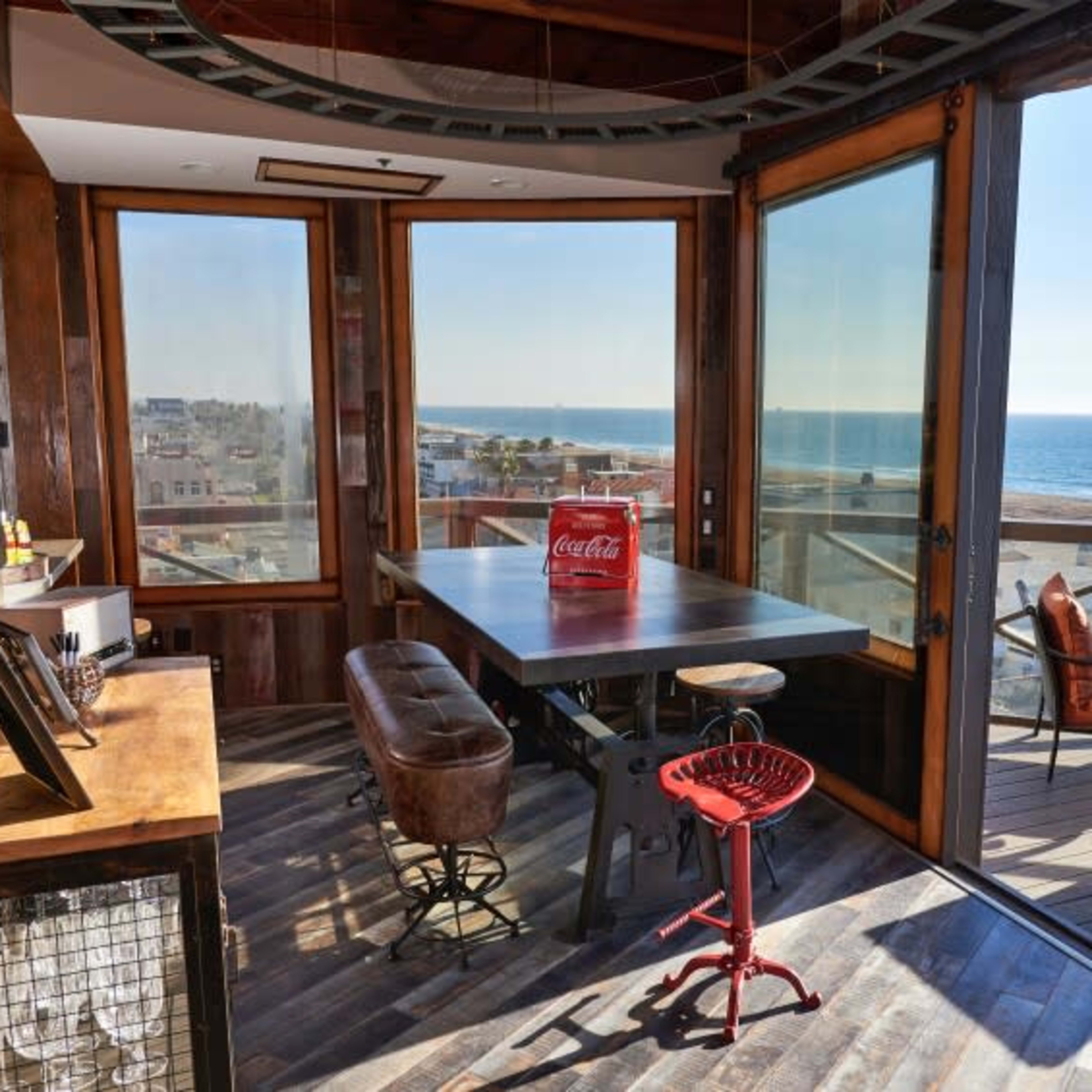 A spacious room with large windows overlooks the ocean, featuring a wooden table and chairs, creating a bright and open atmosphere.
