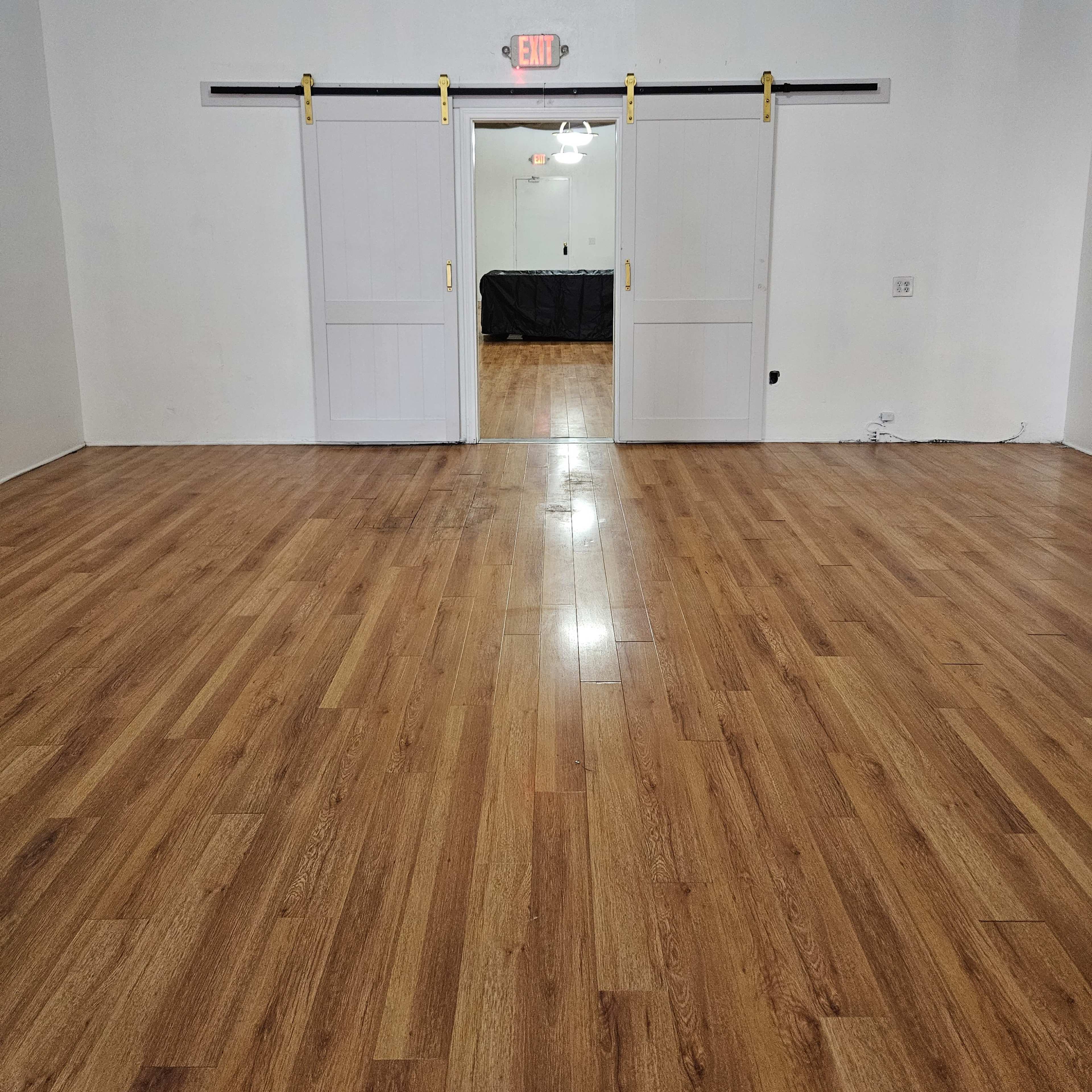 A room with light wooden flooring and two sliding barn doors in the center, leading to another room with an exit sign above.