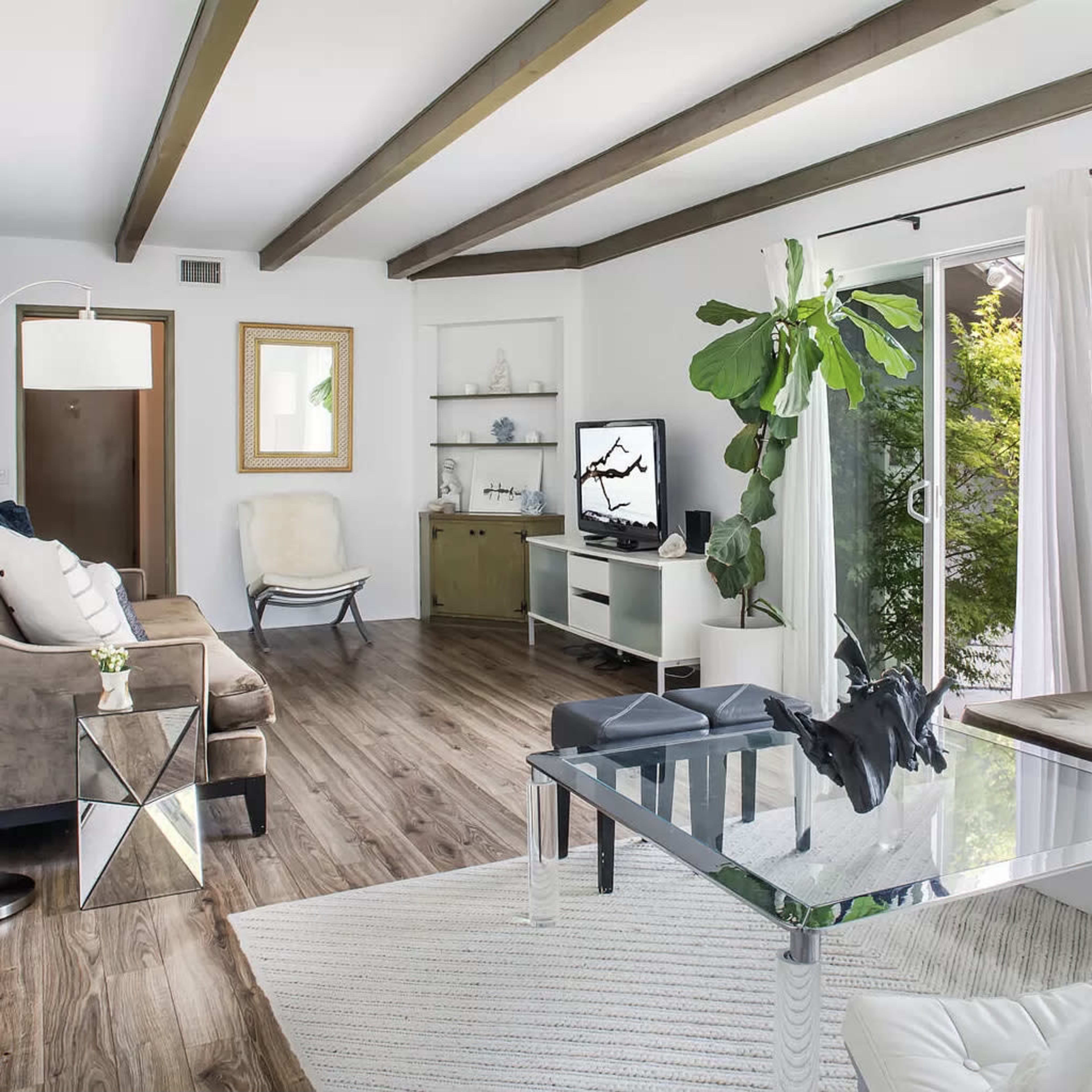 A modern living room with wooden beams on the ceiling, a glass coffee table, and large windows allowing natural light to enter.