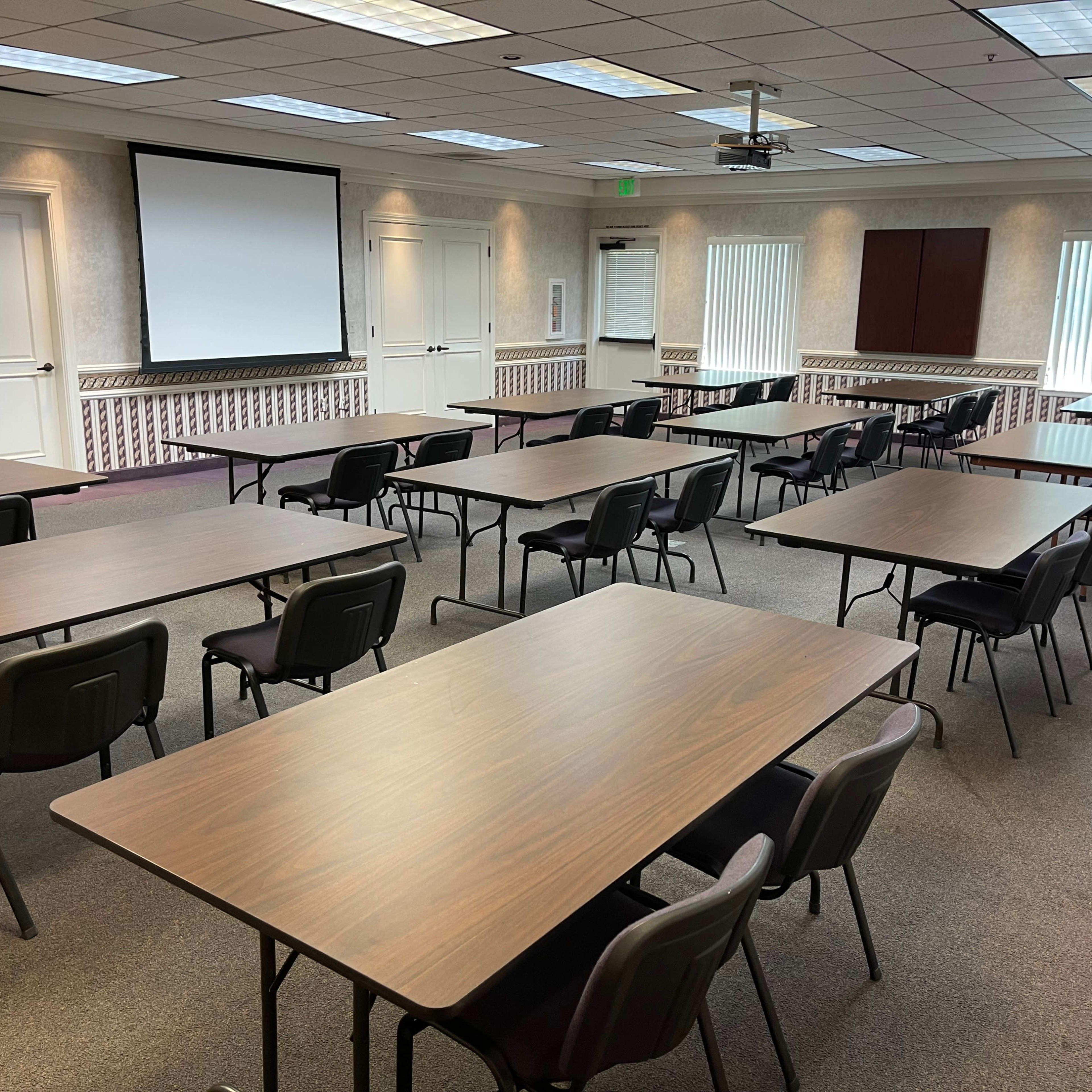 A conference room arranged with multiple tables and chairs, facing a projection screen and windows.