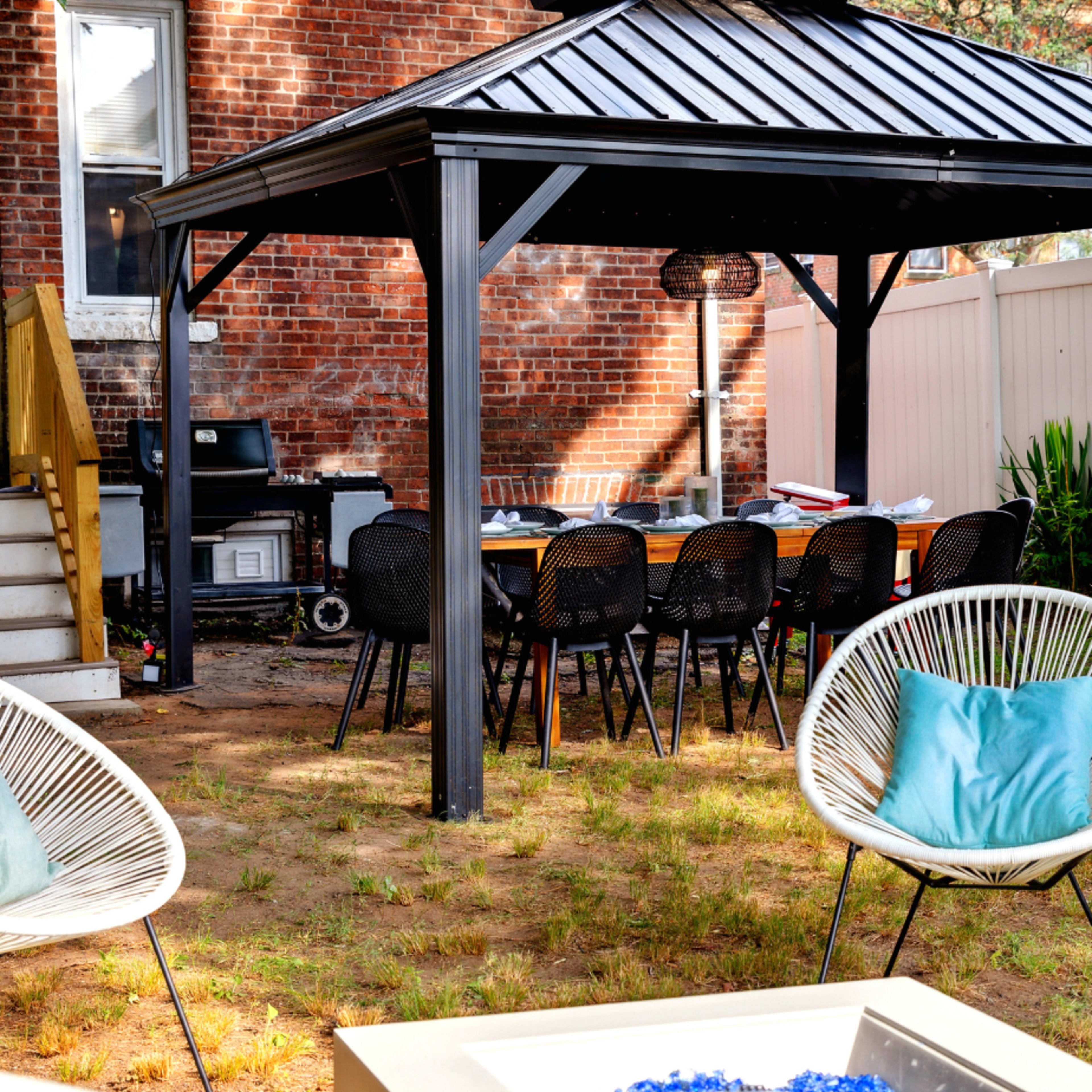 A backyard features a black gazebo with a long table and chairs underneath, surrounded by a grassy area and a seating arrangement with white chairs.
