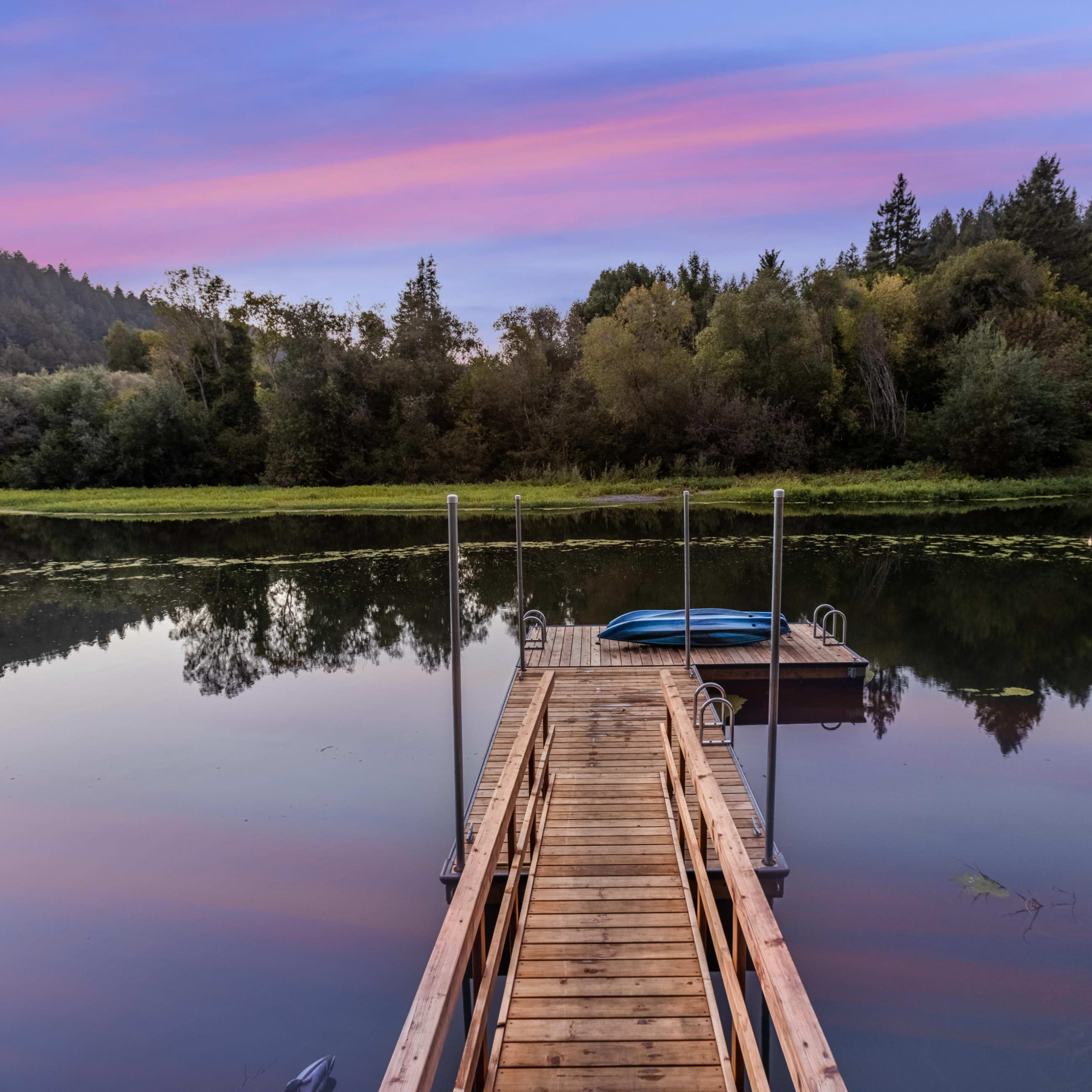 A wooden dock extends into a calm lake surrounded by trees, with a blue kayak resting nearby.