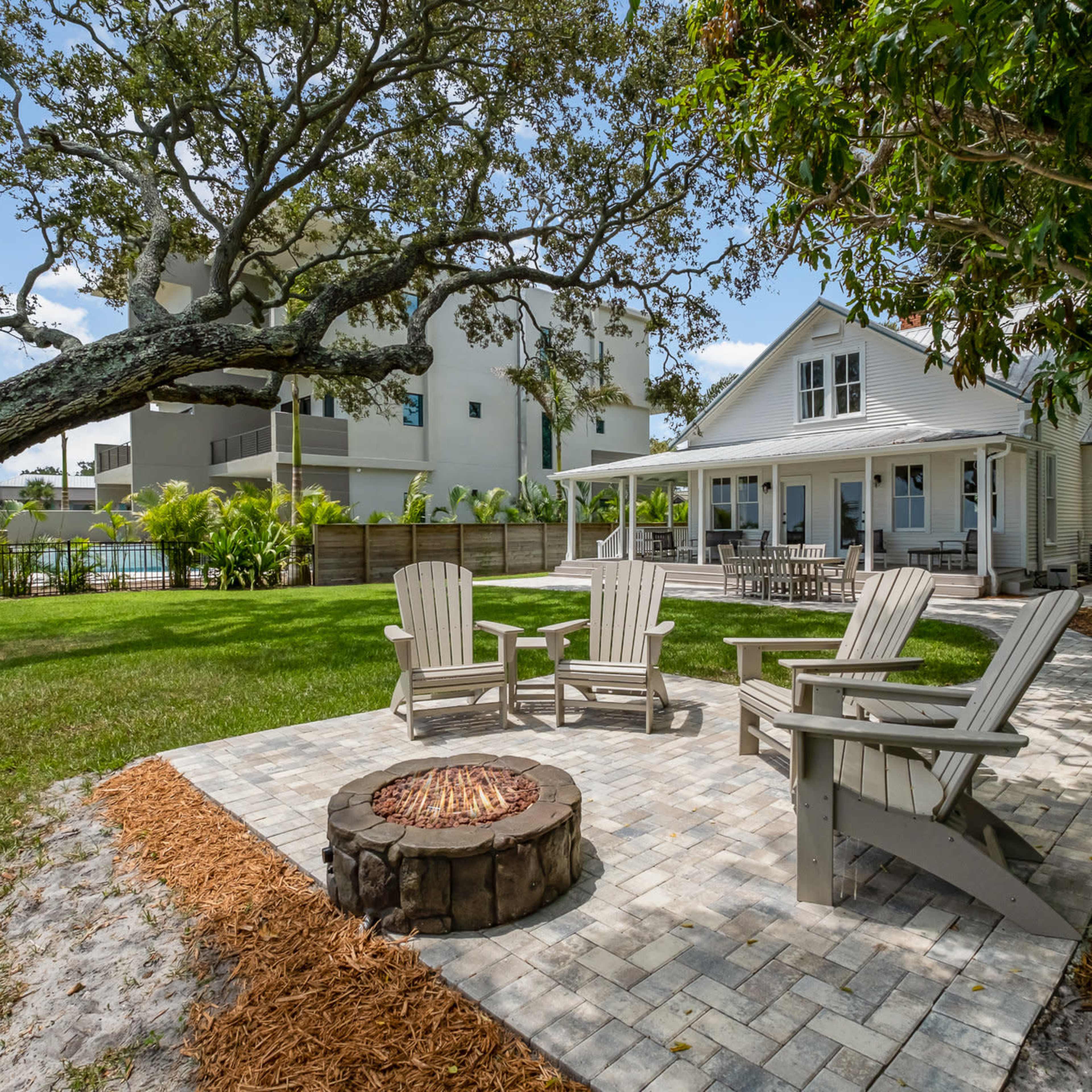A backyard features a circular stone patio with four Adirondack chairs arranged around a fire pit, surrounded by lush greenery and a large tree, with a house and a high-rise building in the background.