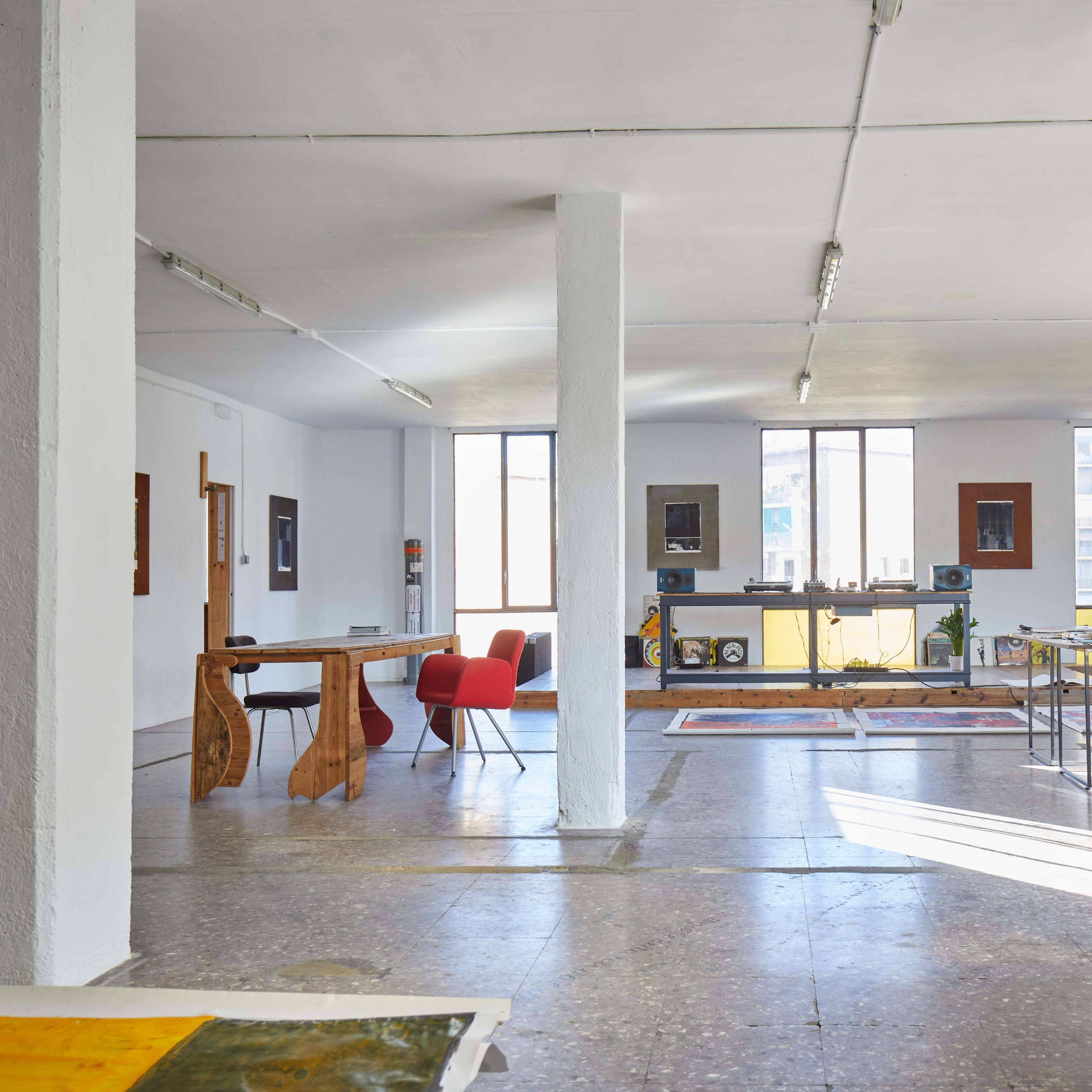 A spacious, minimalist art studio with large windows, featuring a white sofa, a wooden table, and various artworks displayed on the walls.