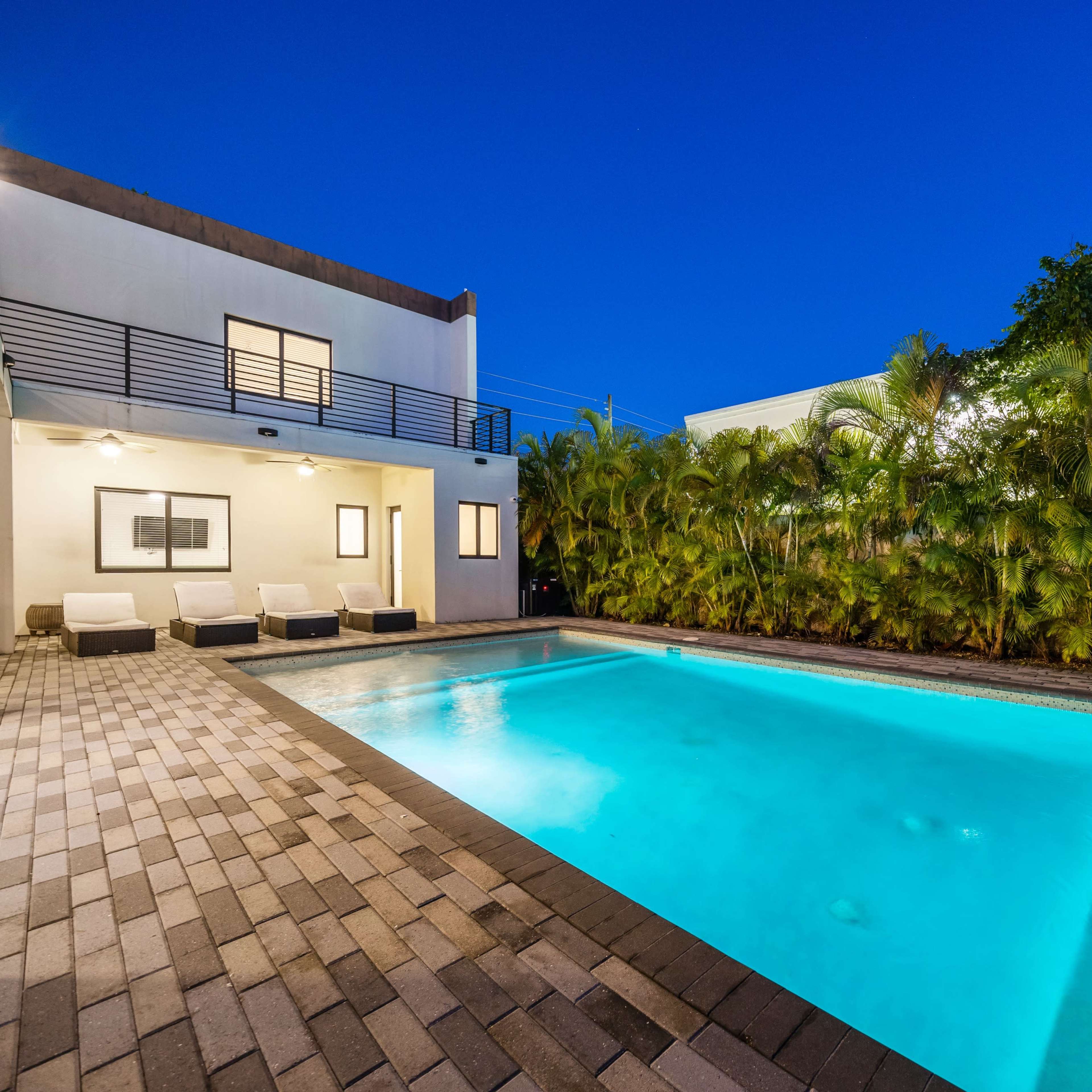 A modern backyard with a swimming pool surrounded by patio pavers and tropical plants under a twilight sky.