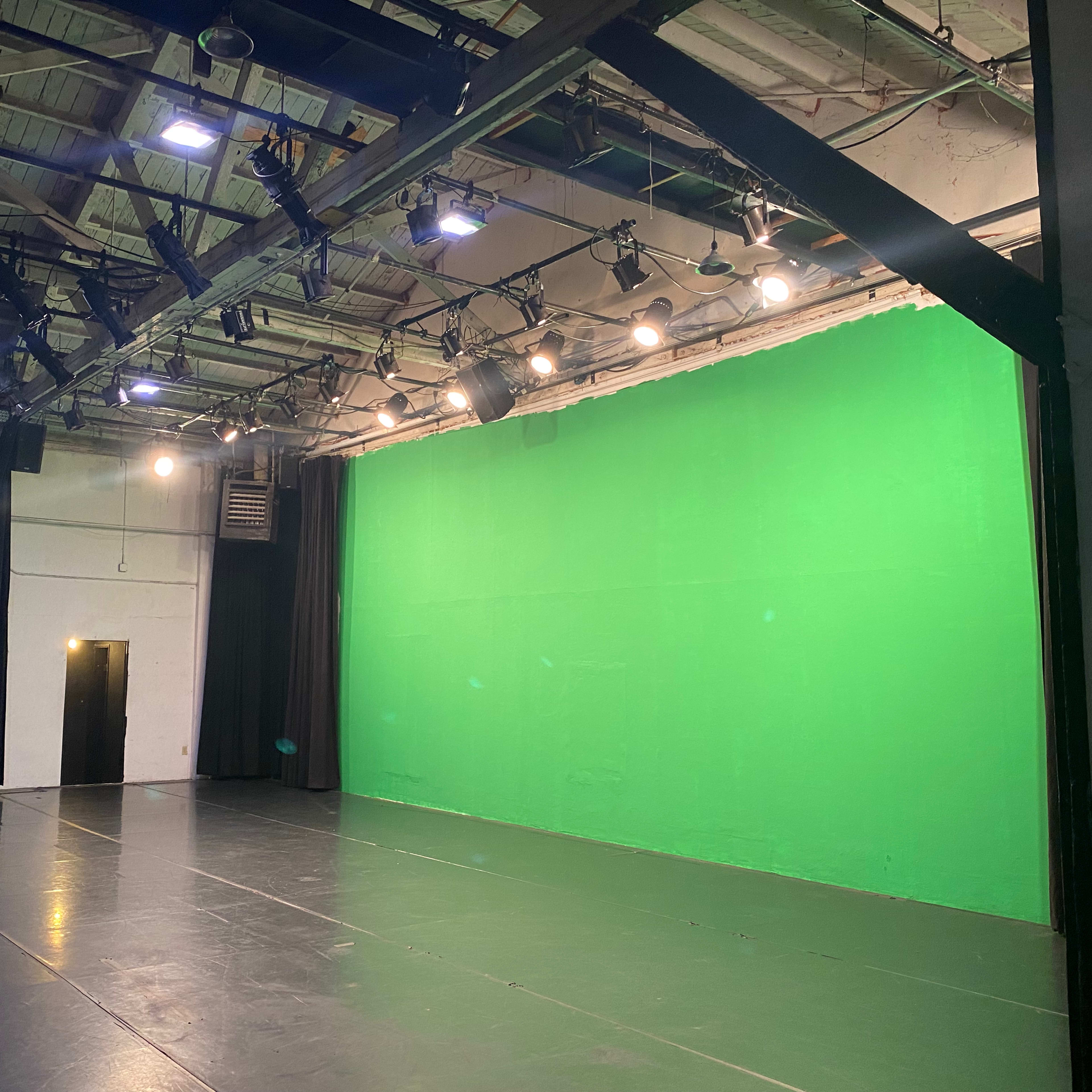 An empty studio space with a bright green screen on one wall and overhead lighting fixtures.