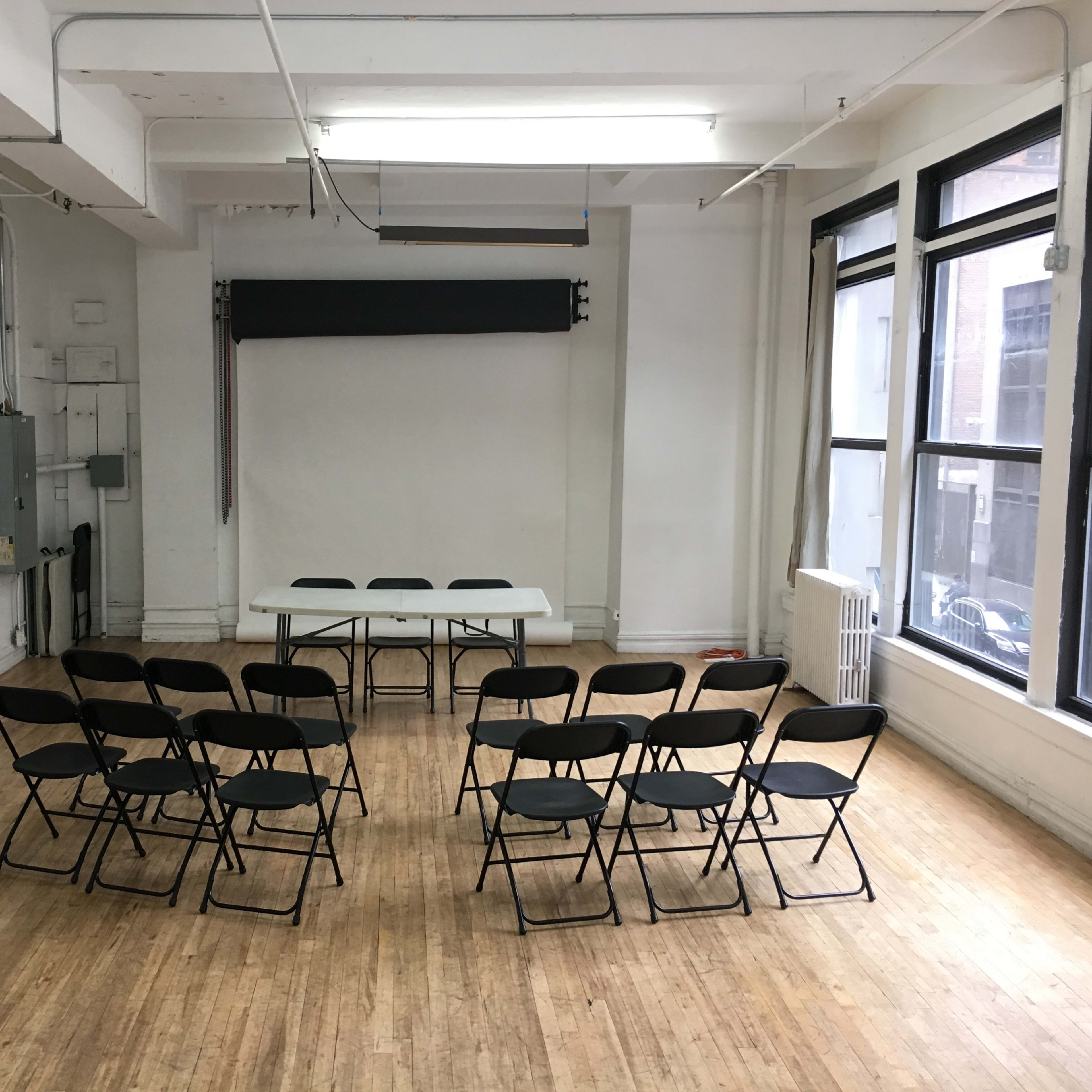 A simple room with a white backdrop and a long table surrounded by folding chairs arranged in a semicircle.