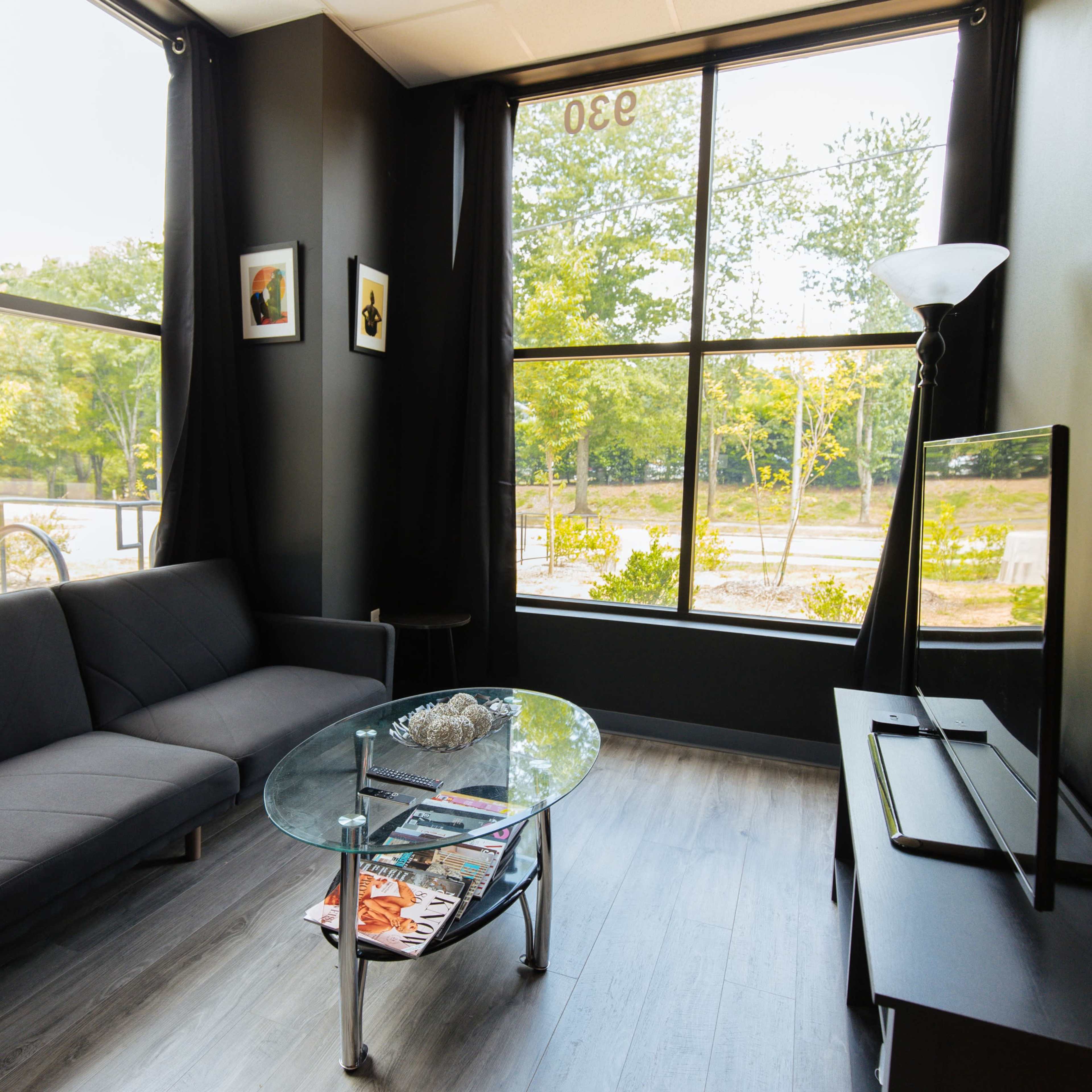 A modern living room features a black sofa and a glass coffee table, with large windows providing a view of greenery outside.