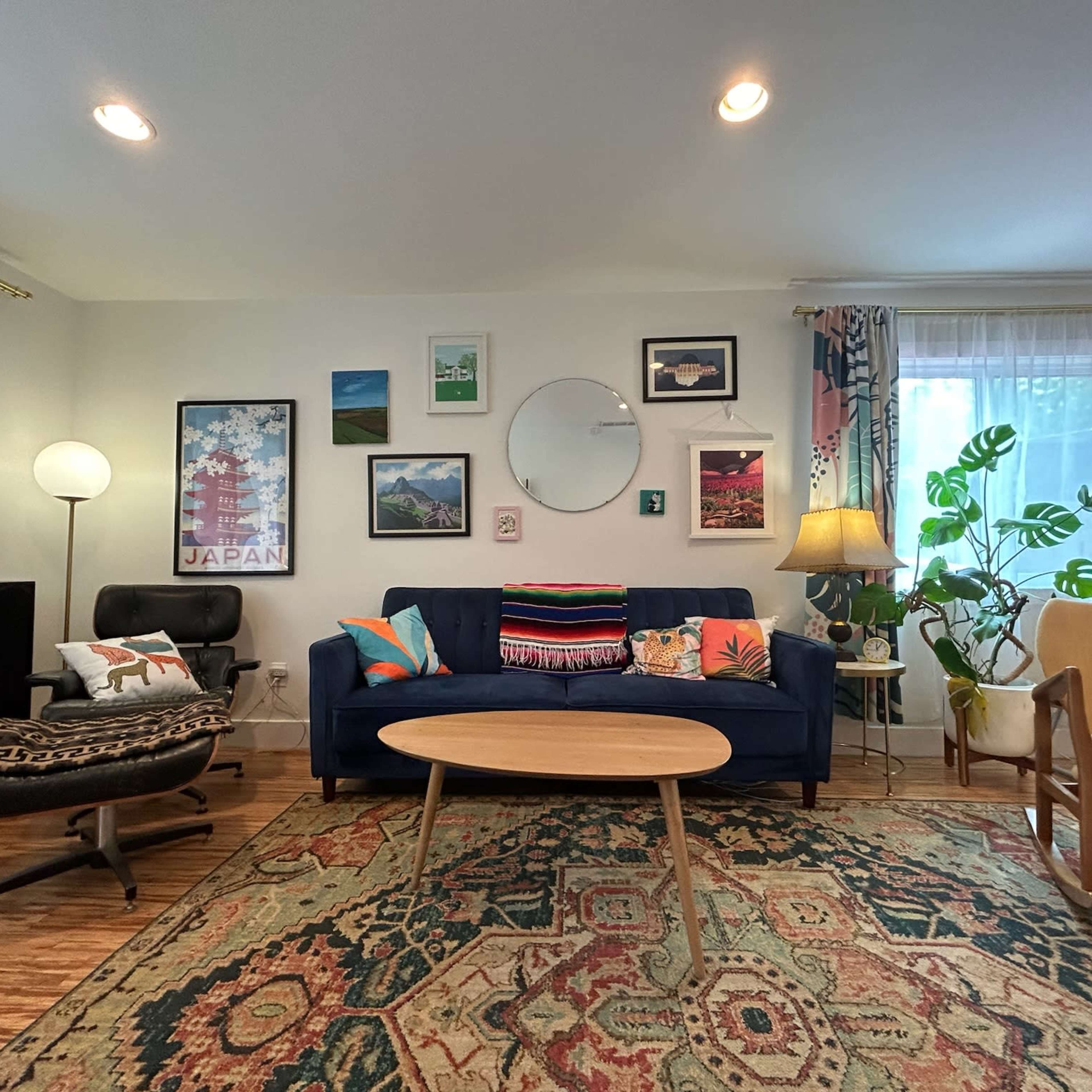 The living room features a blue couch with assorted cushions, a round coffee table, and various framed artworks on the walls, complemented by plants and additional seating.