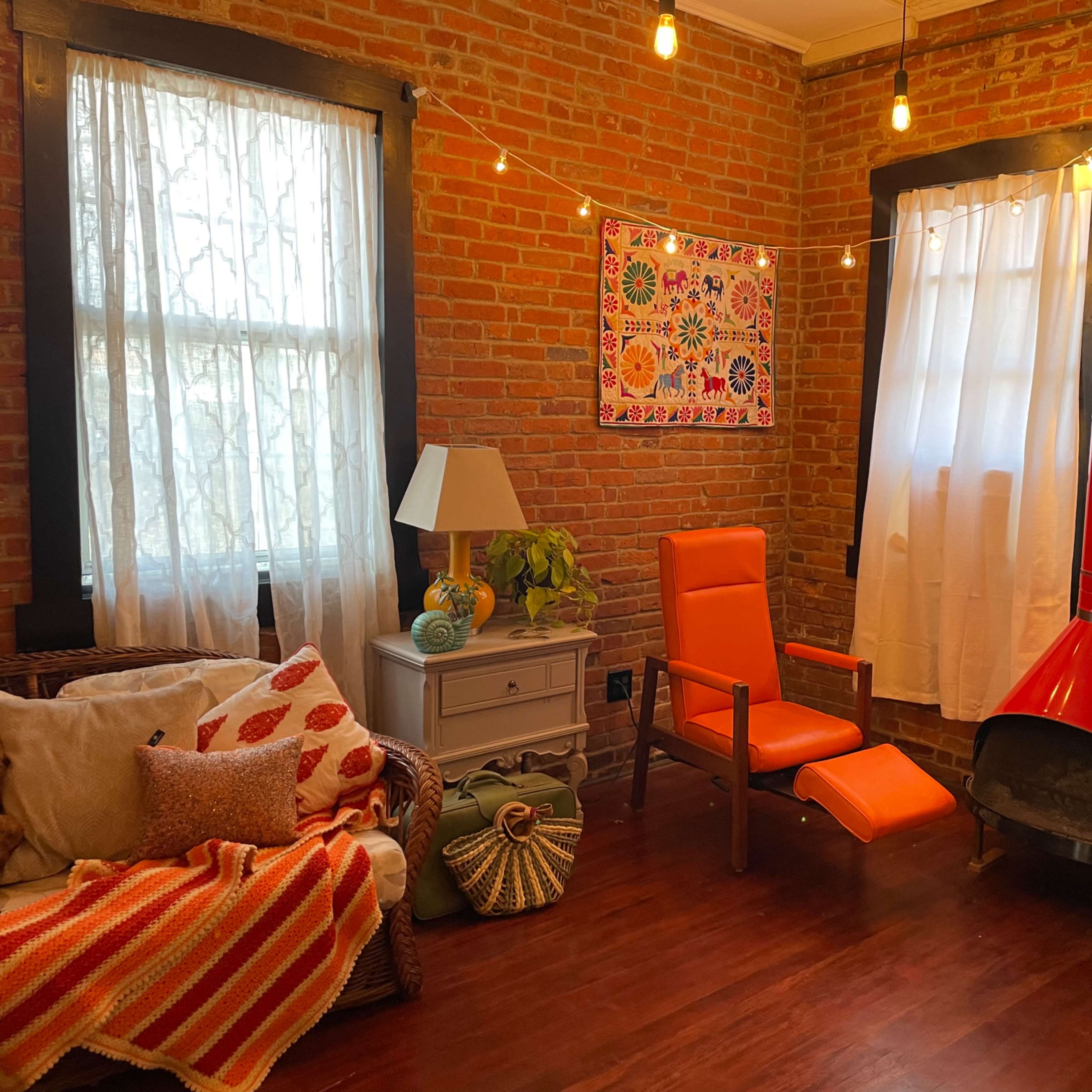 A cozy living room featuring a brick wall, a red-painted fireplace, a bright orange chair, and a decorative lamp beside a sofa with patterned cushions.
