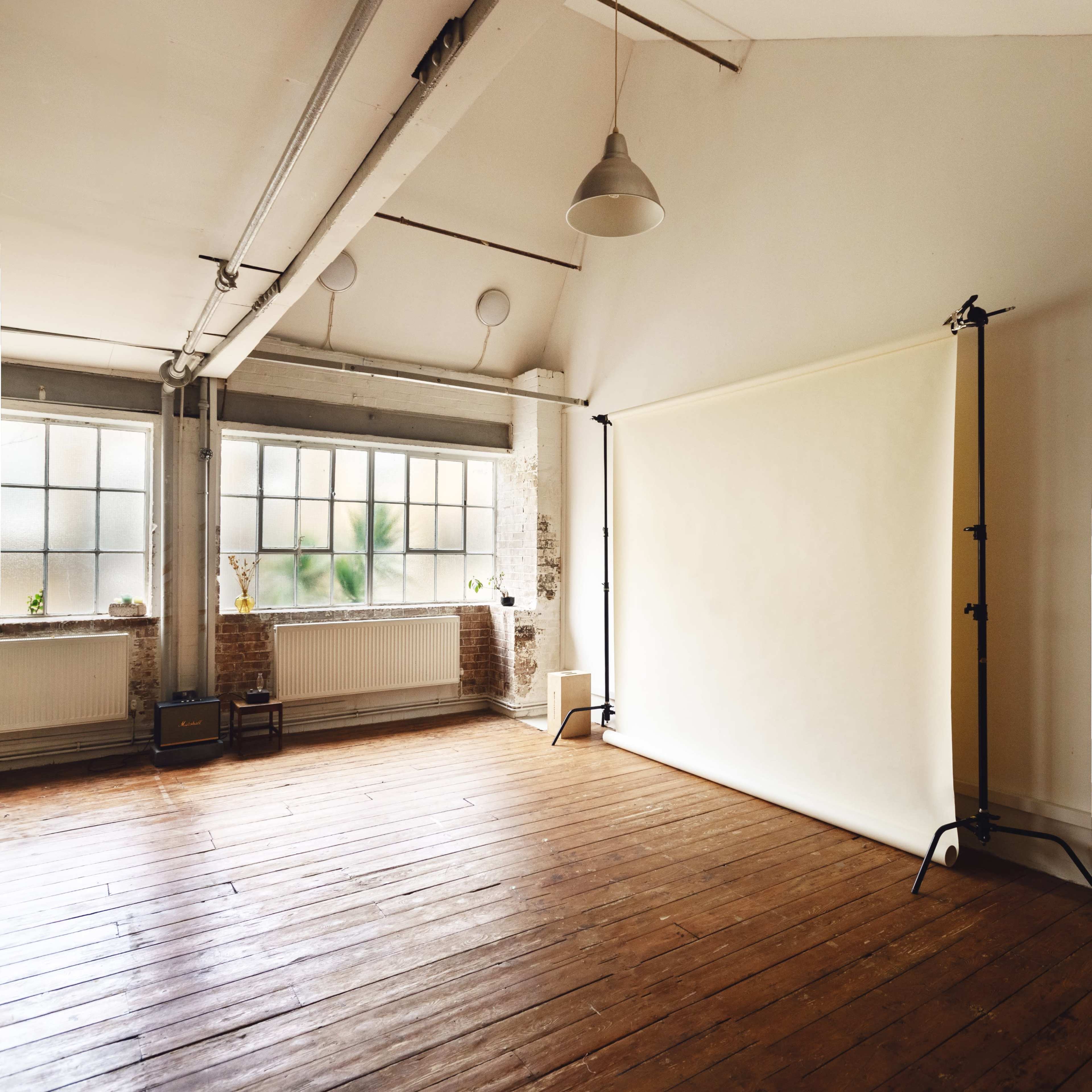 A spacious, well-lit studio with wooden floors, large windows, and a plain backdrop set up for photography.