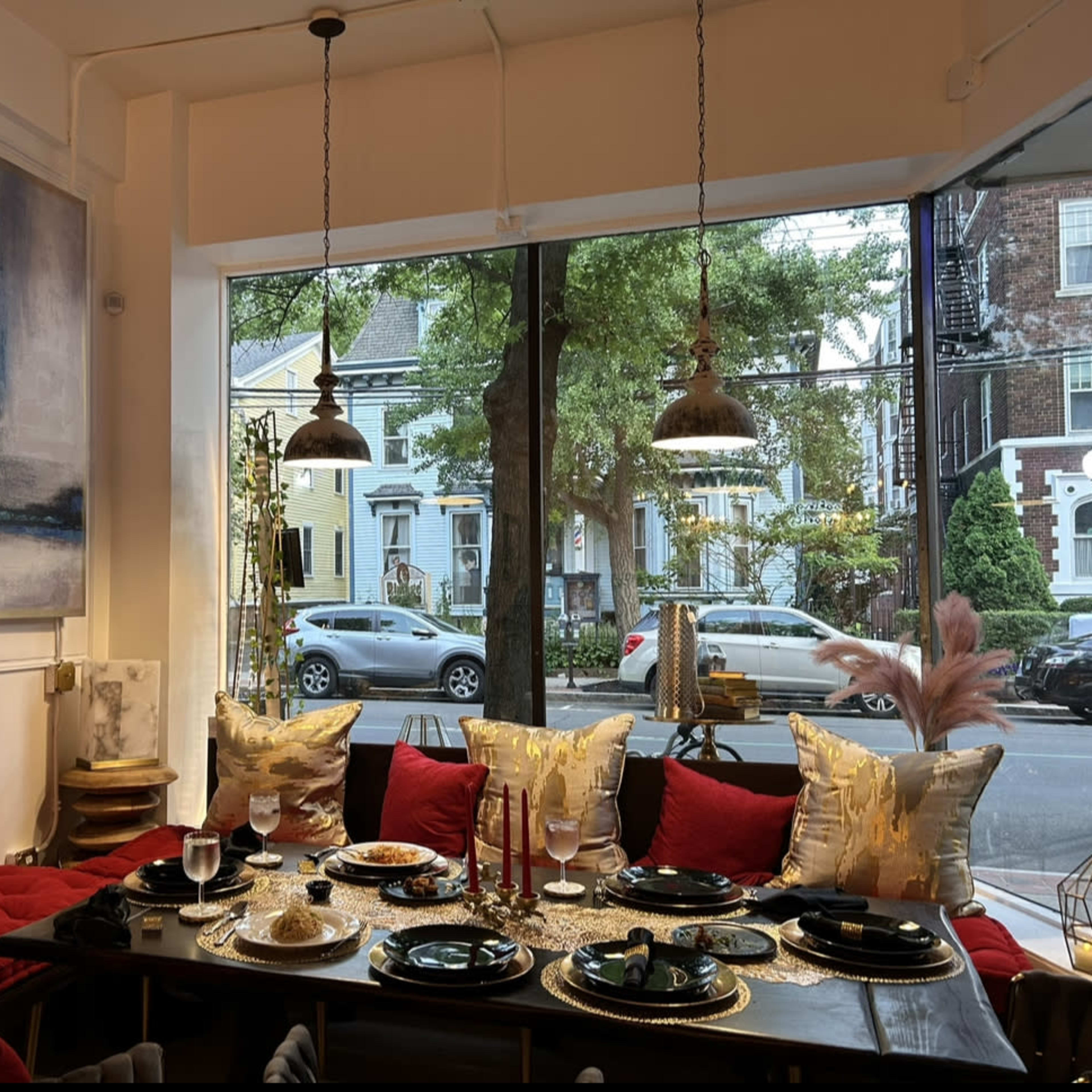 A dining area features a table set with elegant dishes, surrounded by cushions and adorned with artistic lighting, overlooking a street lined with trees and houses.