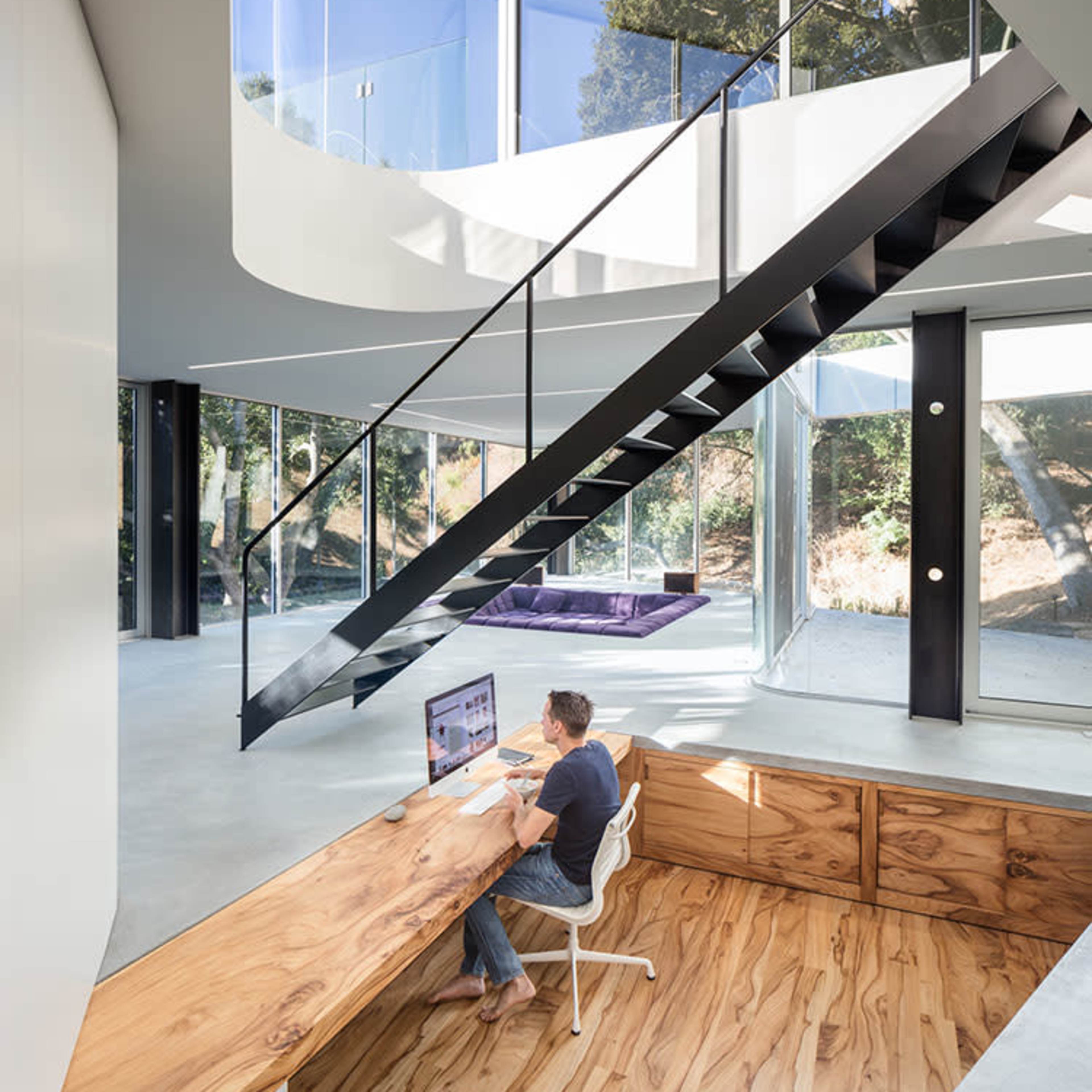 A person works at a minimalist wooden desk in a contemporary home with large windows and a spiral staircase.