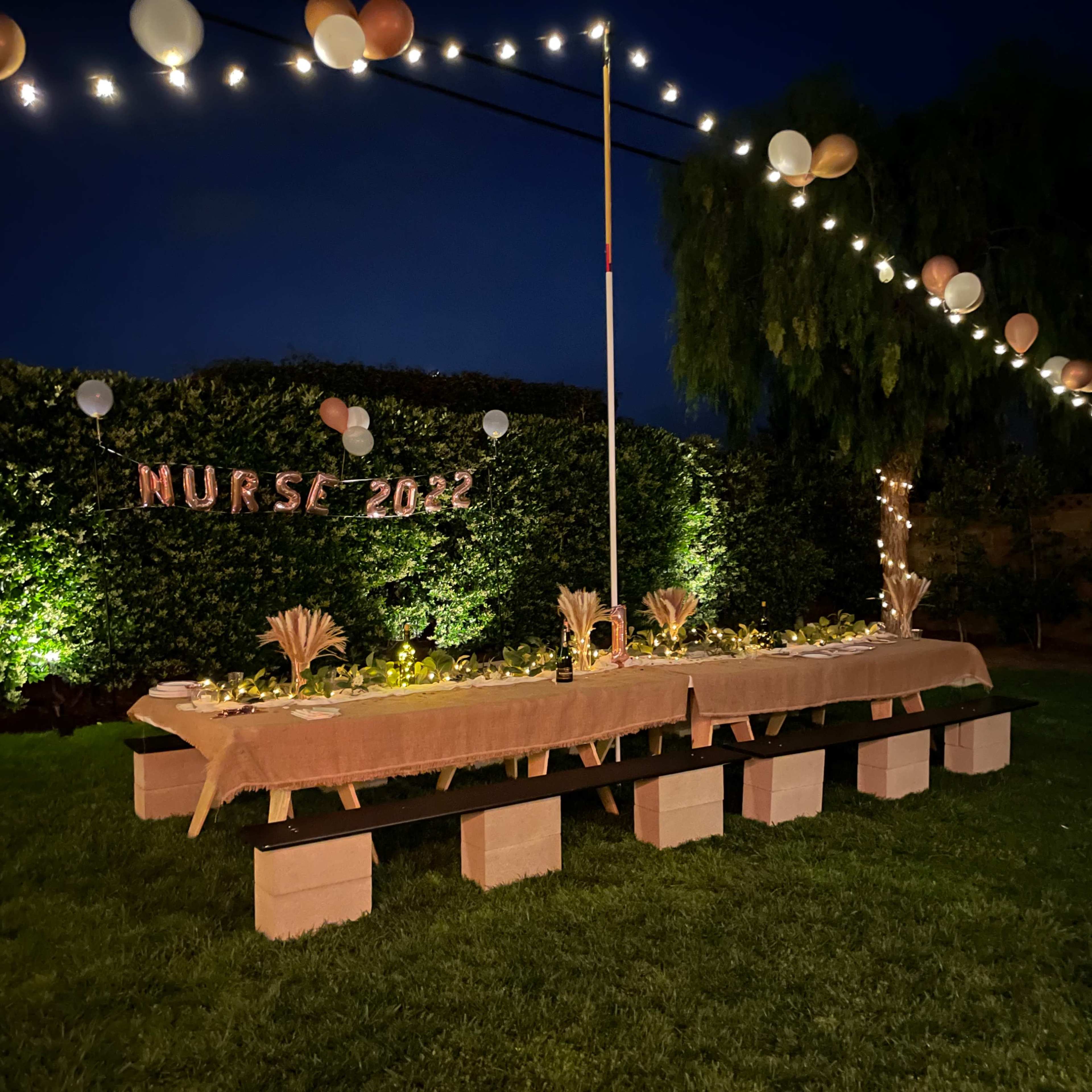 A long table is set with decorations and illuminated by string lights in a garden area for a celebration labeled "NURSE 2022."