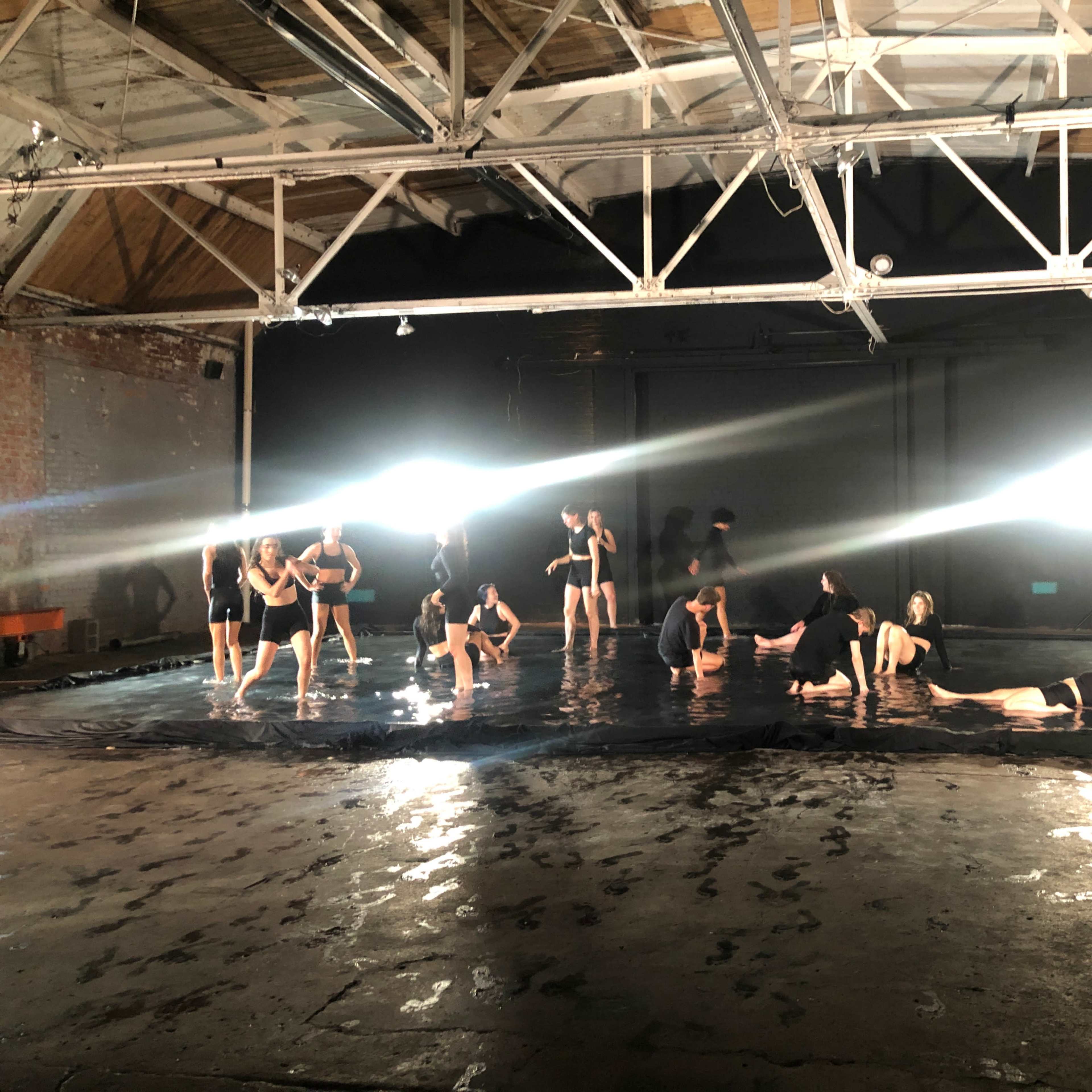 A group of individuals performs on a large black tarp in a dimly lit industrial space.