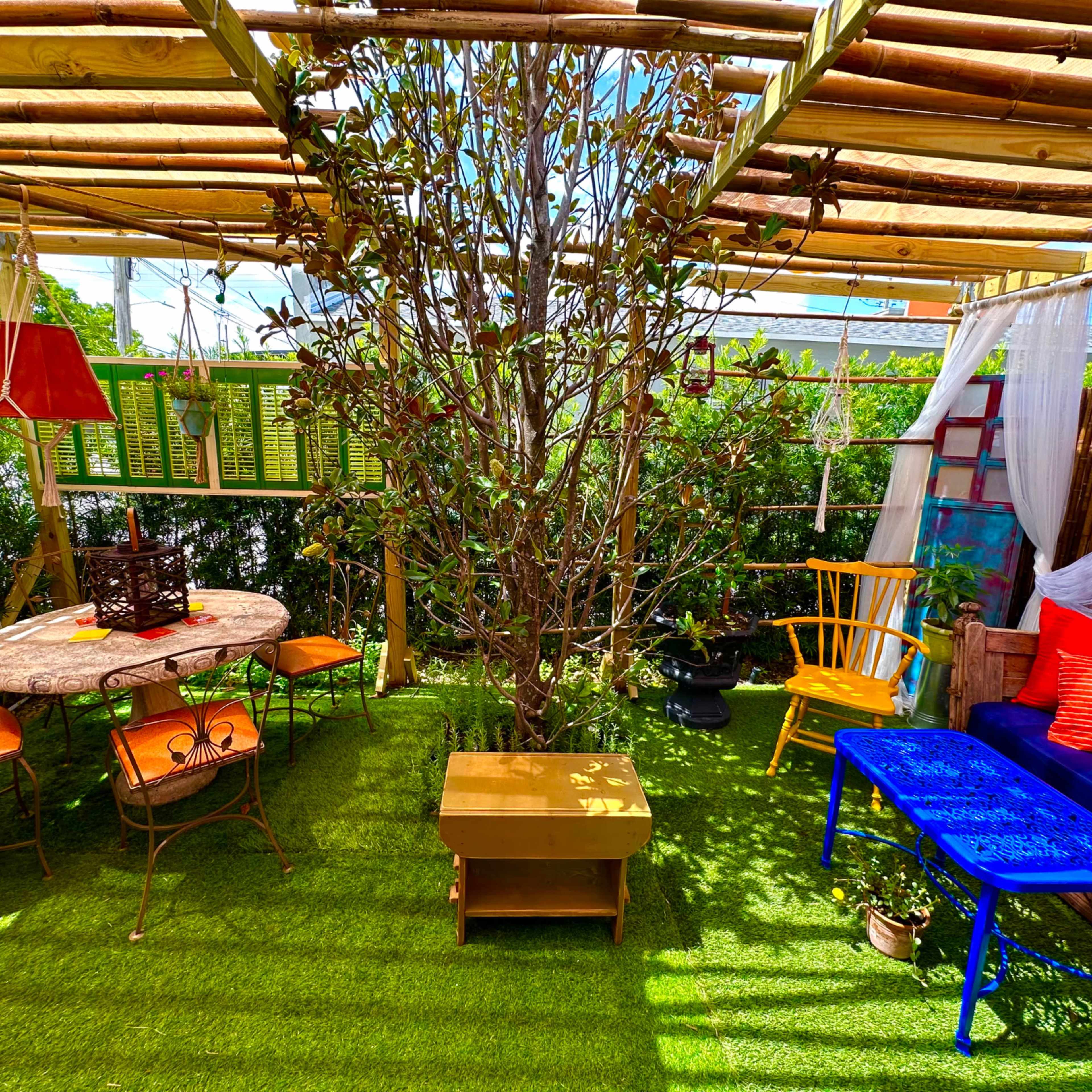 A vibrant outdoor seating area with a table, chairs, a tree, and colorful decor under a bamboo canopy.