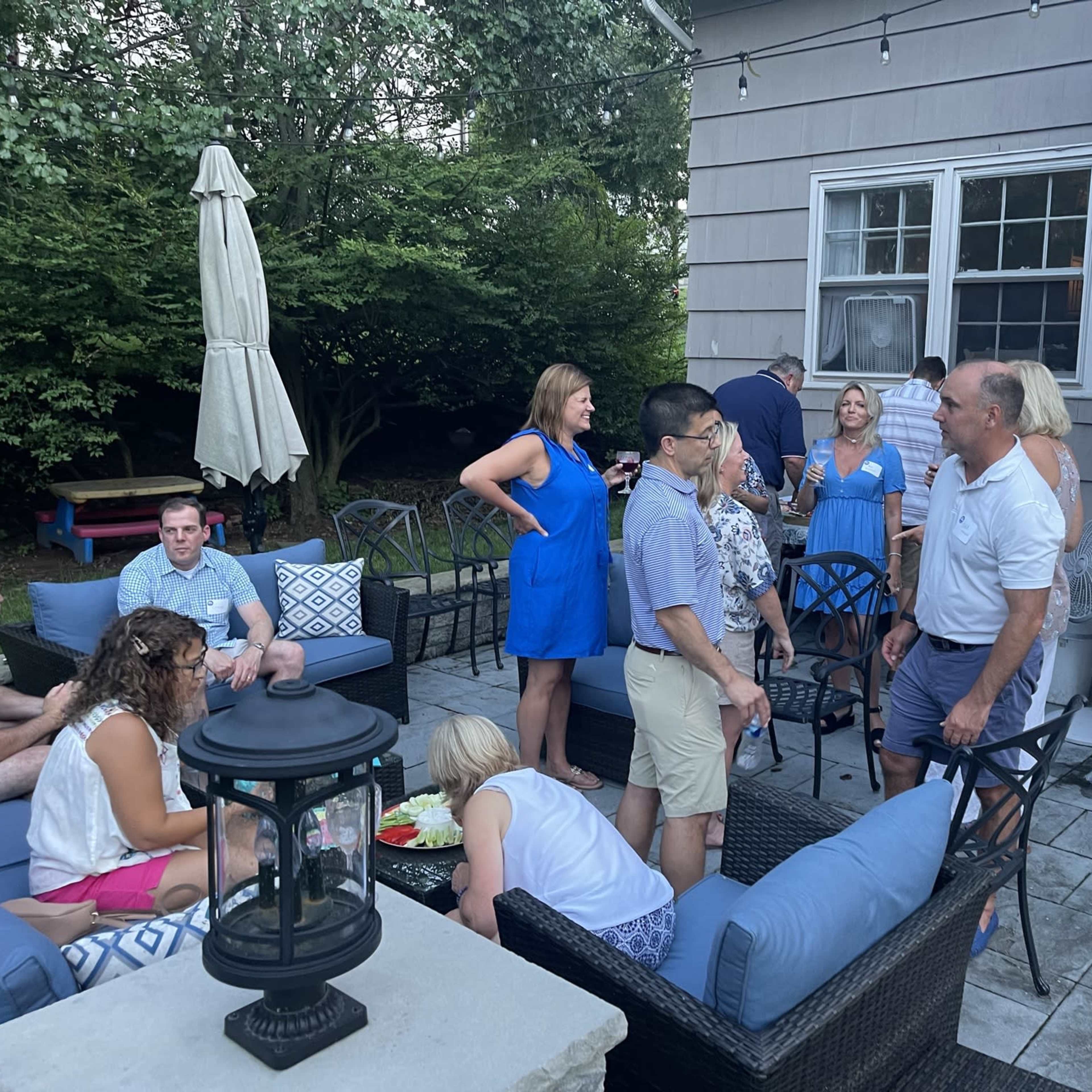 A group of people gather in a backyard setting, engaging in conversation around patio furniture.