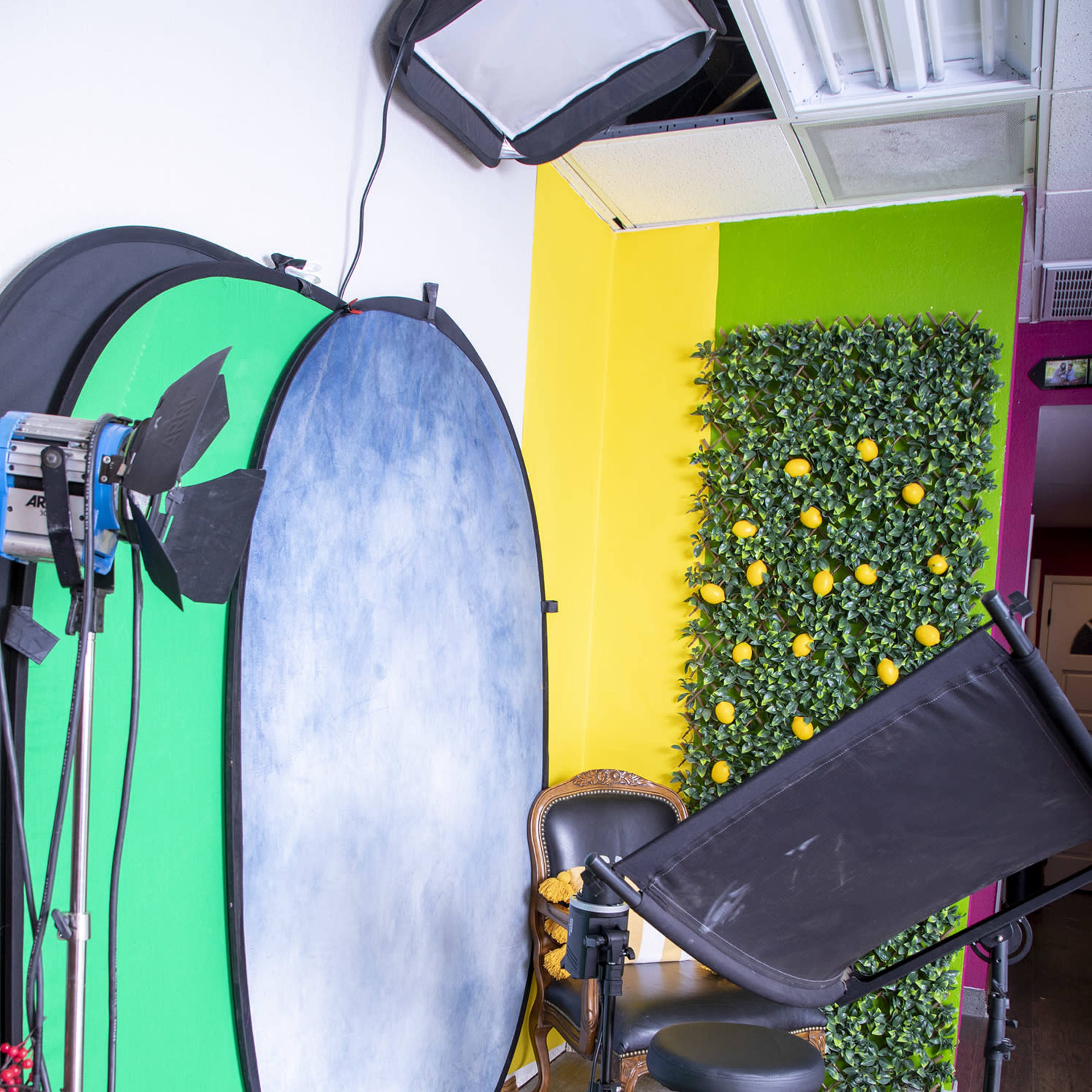 A photography studio setup featuring a green screen, a backdrop, lighting equipment, and a chair placed in front of a decorative wall with faux greenery and yellow accents.