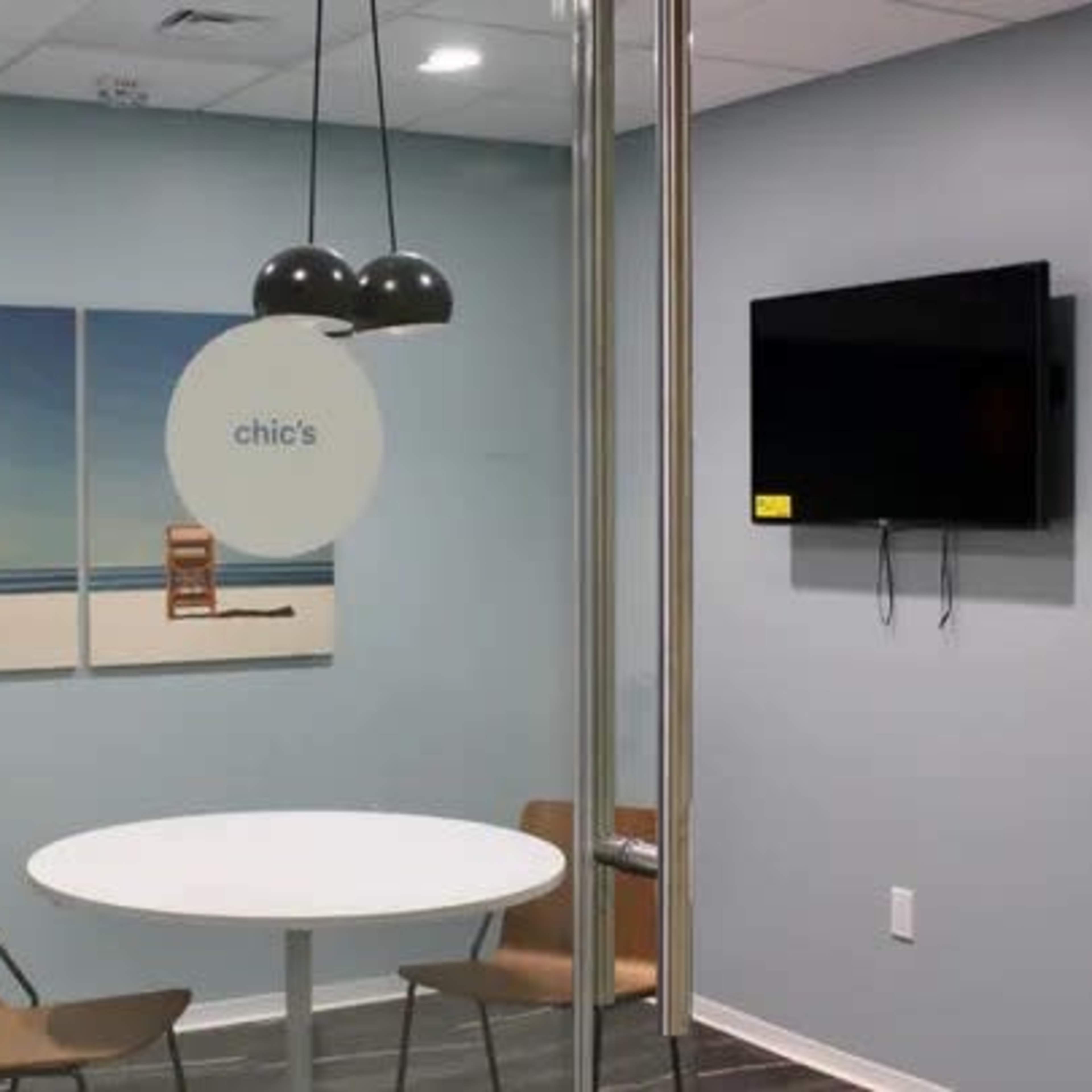 A modern glass-walled meeting room featuring a round table, two chairs, a TV, and beach-themed artwork on the wall.
