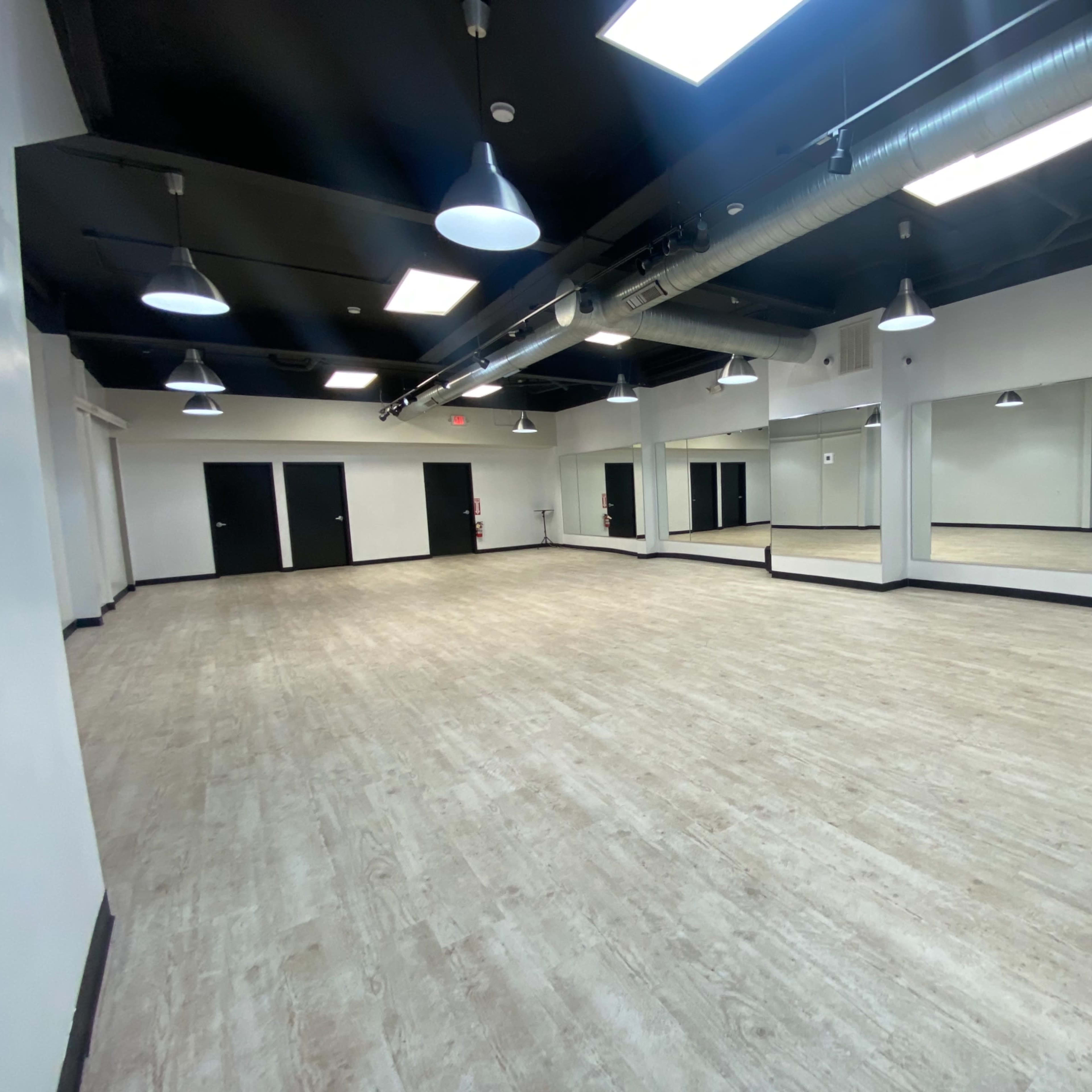 A spacious, empty room with wooden floors, mirrored walls, and modern lighting fixtures.