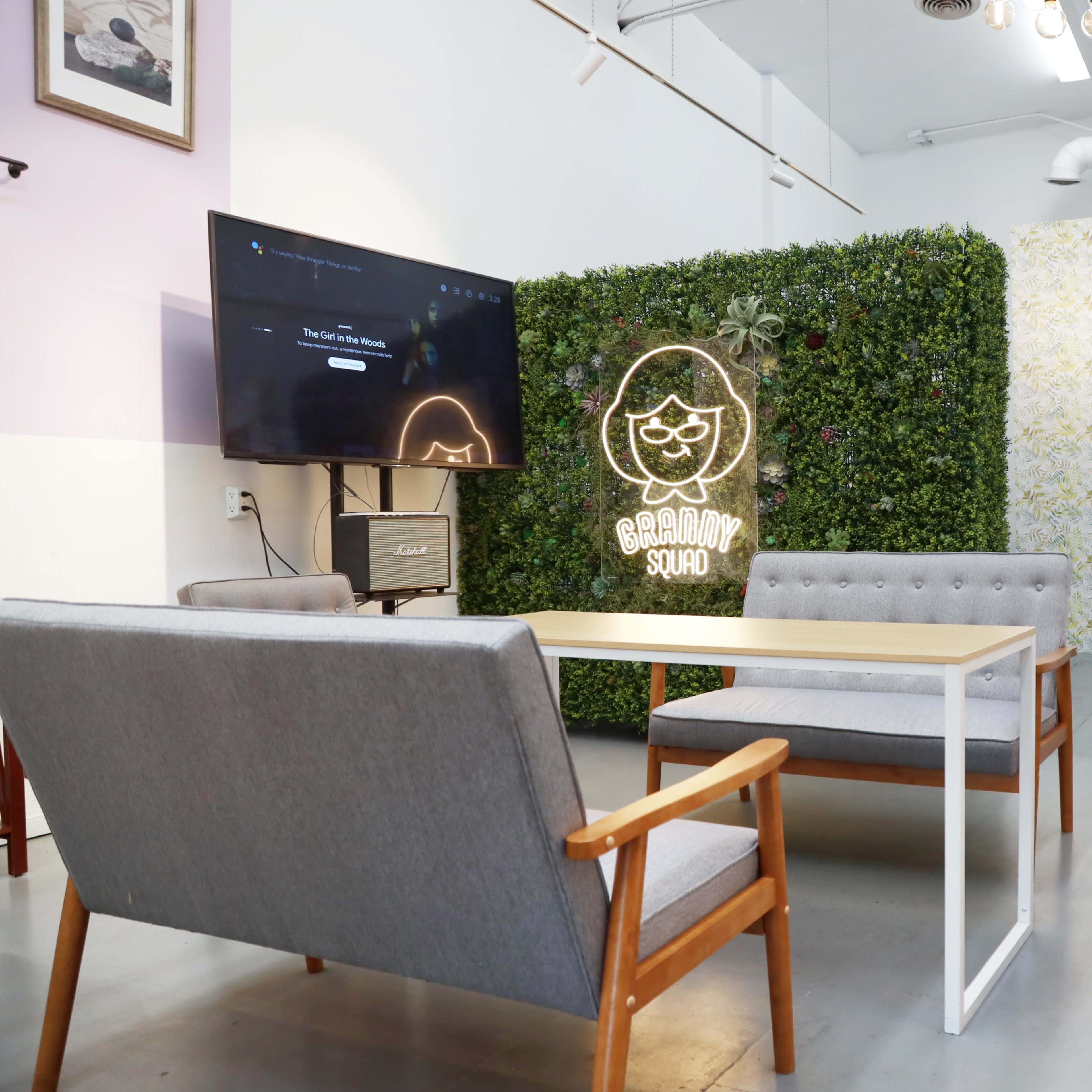A modern lounge space features a wooden table surrounded by four chairs, with a wall of greenery and decorative wall art in the background.