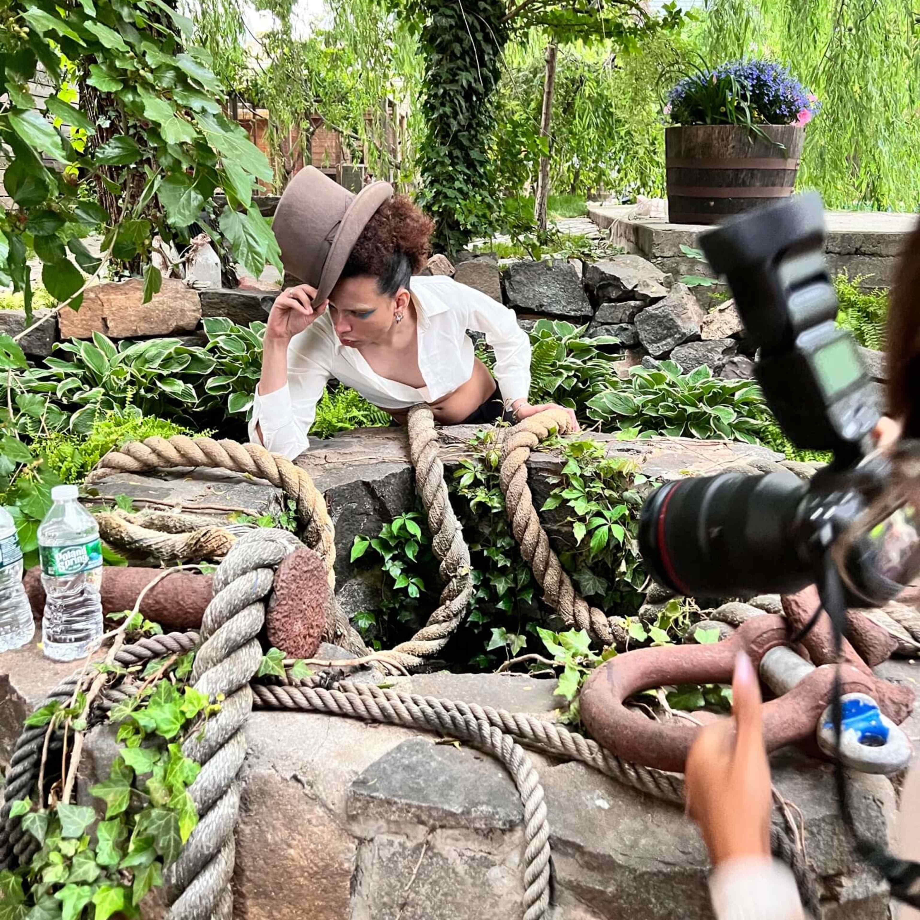 A person is emerging from a stone well surrounded by greenery and old ropes, while another individual nearby is holding a camera.