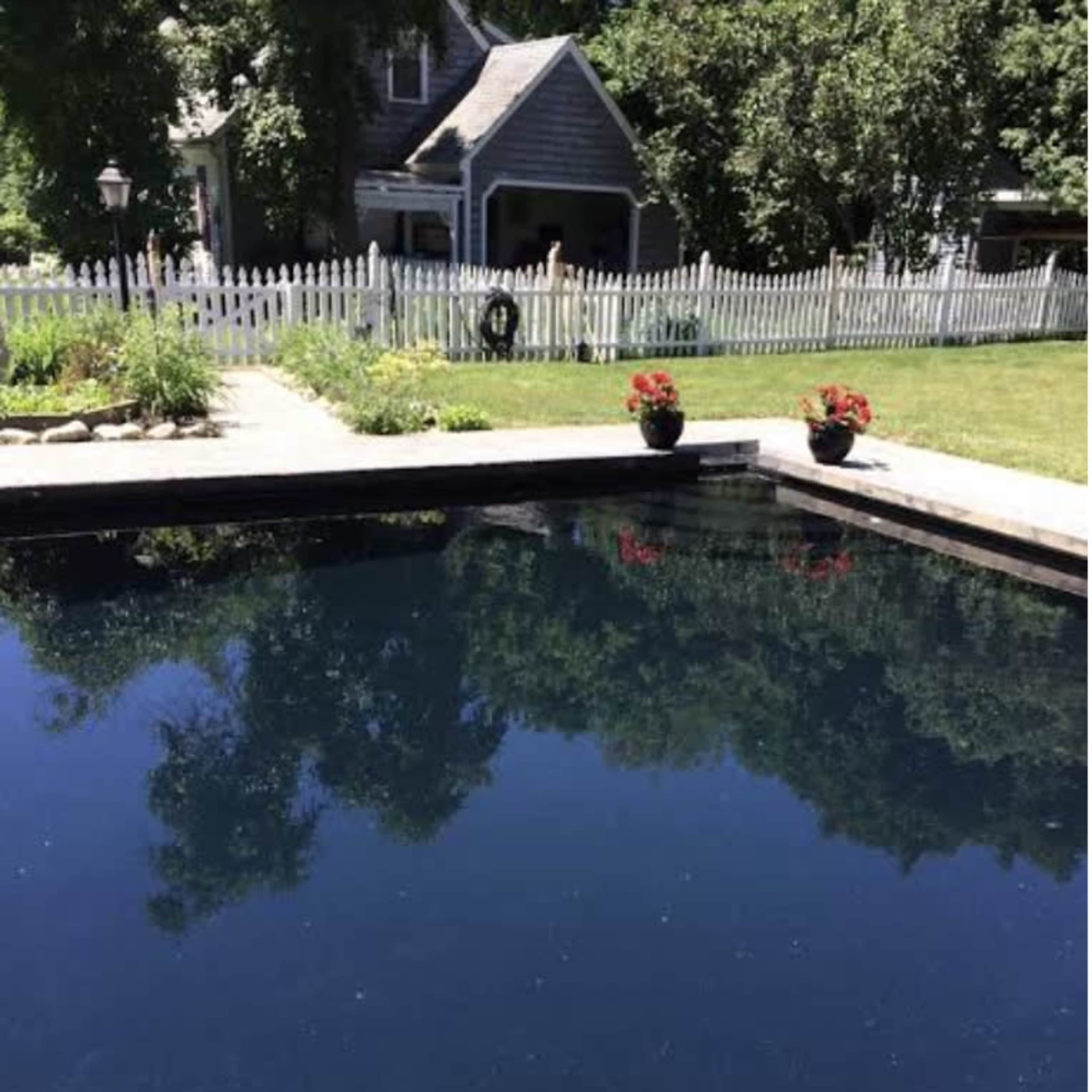A dark, still swimming pool surrounded by a grassy area and a white picket fence, with a house visible in the background.