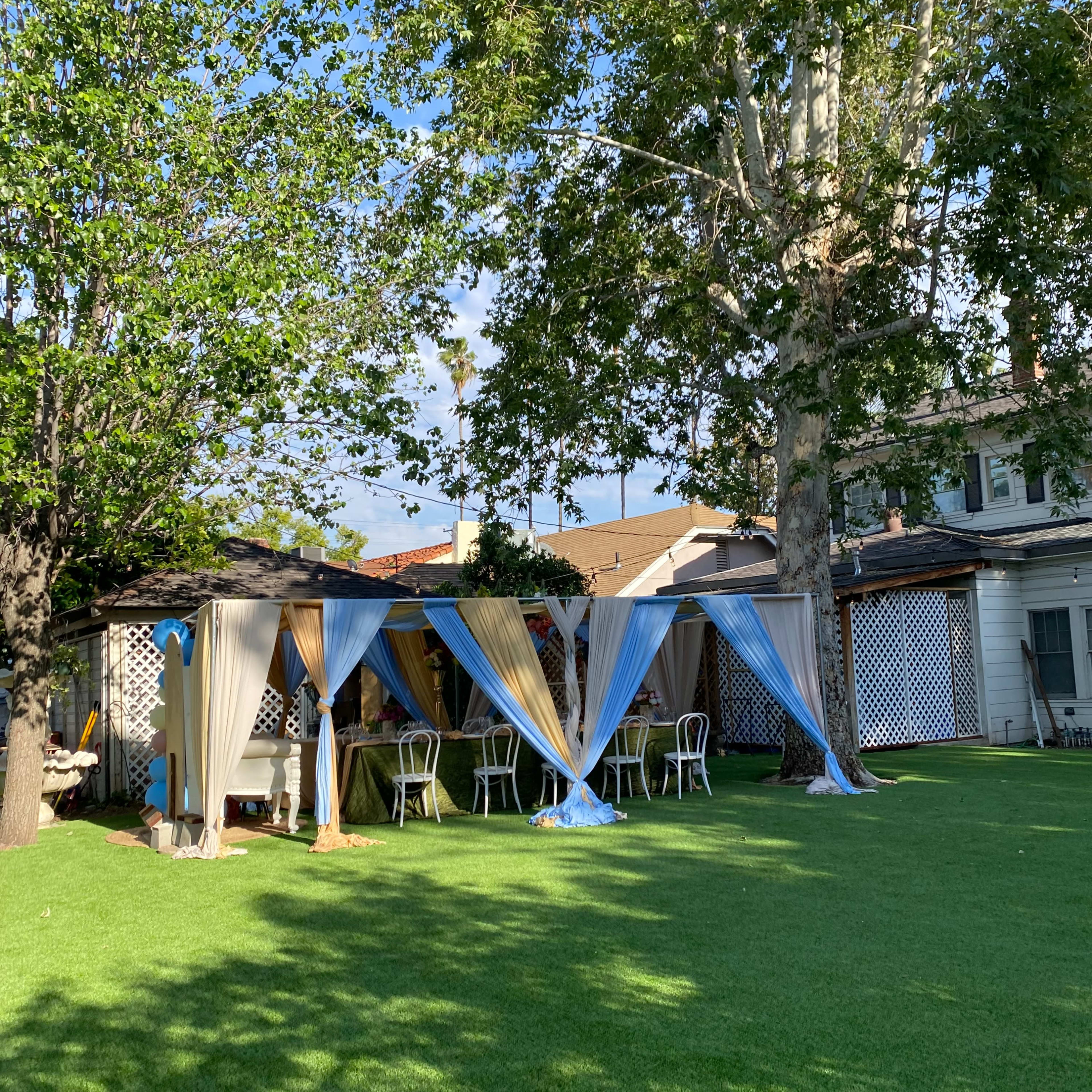 A secluded outdoor area with canopies draped in blue and beige fabric, surrounded by green grass and chairs under a large tree.
