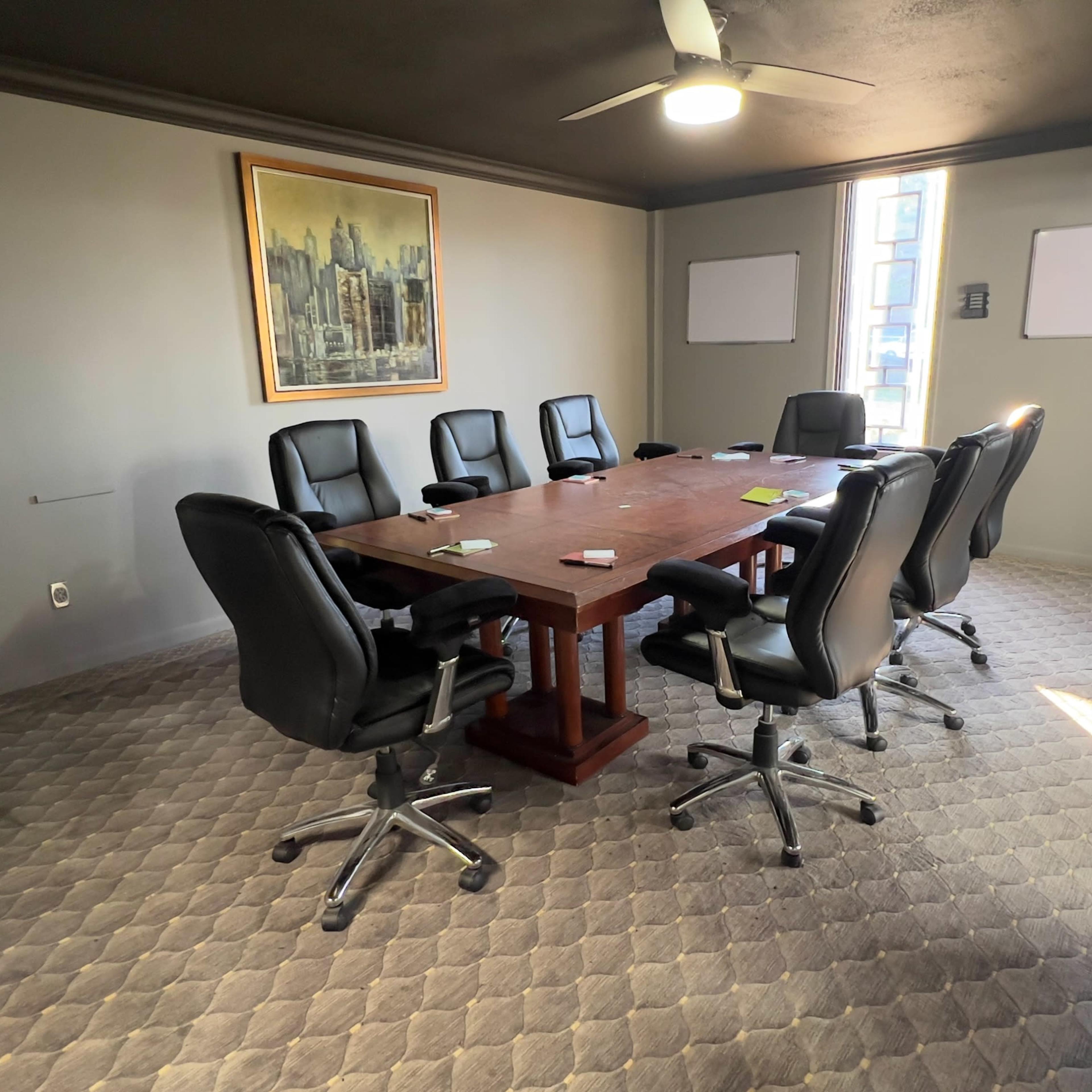 A conference room with a large wooden table surrounded by eight black leather chairs, featuring a painting on the wall and two whiteboards.