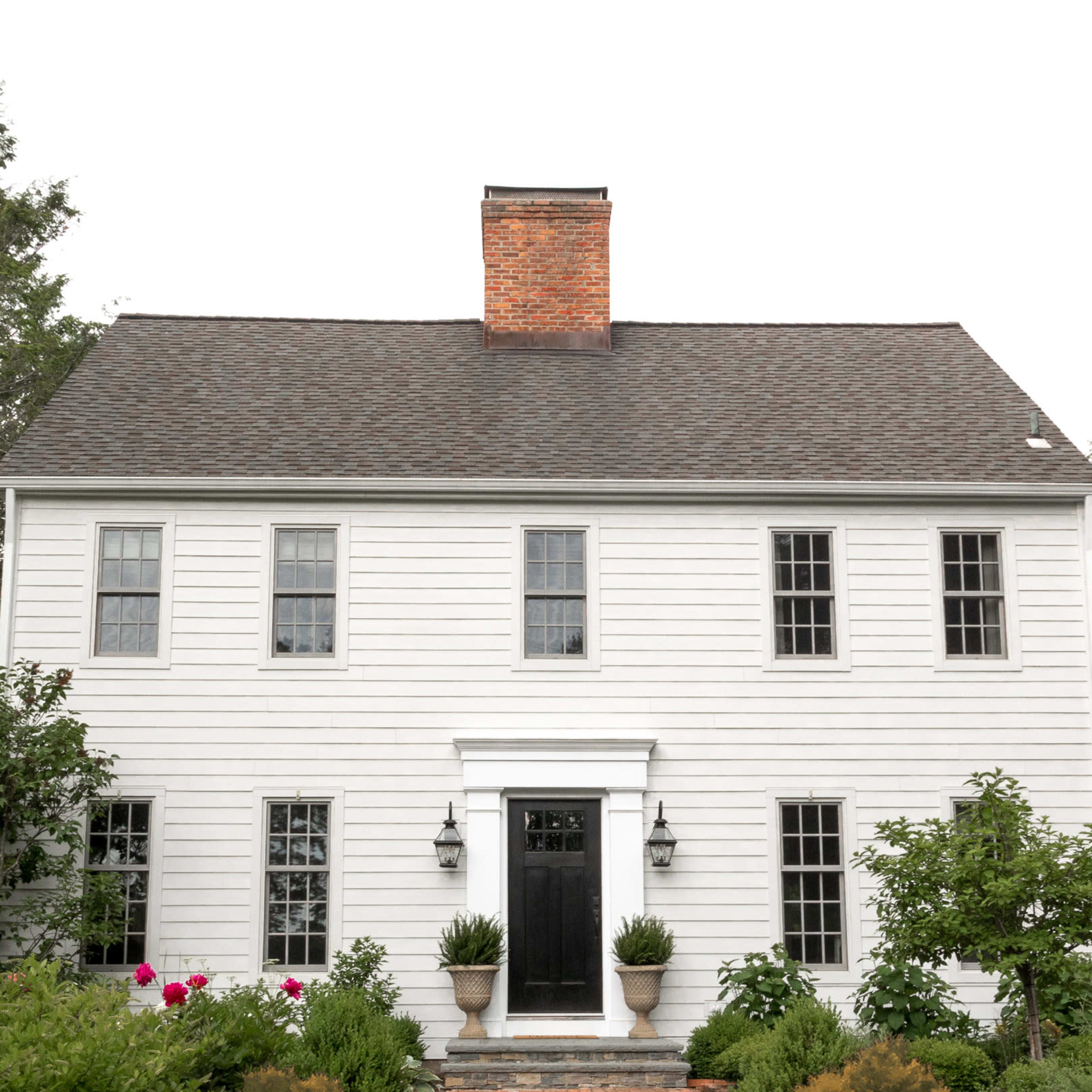 A white wooden house with a central dark door and large windows is surrounded by manicured plants and flowers.