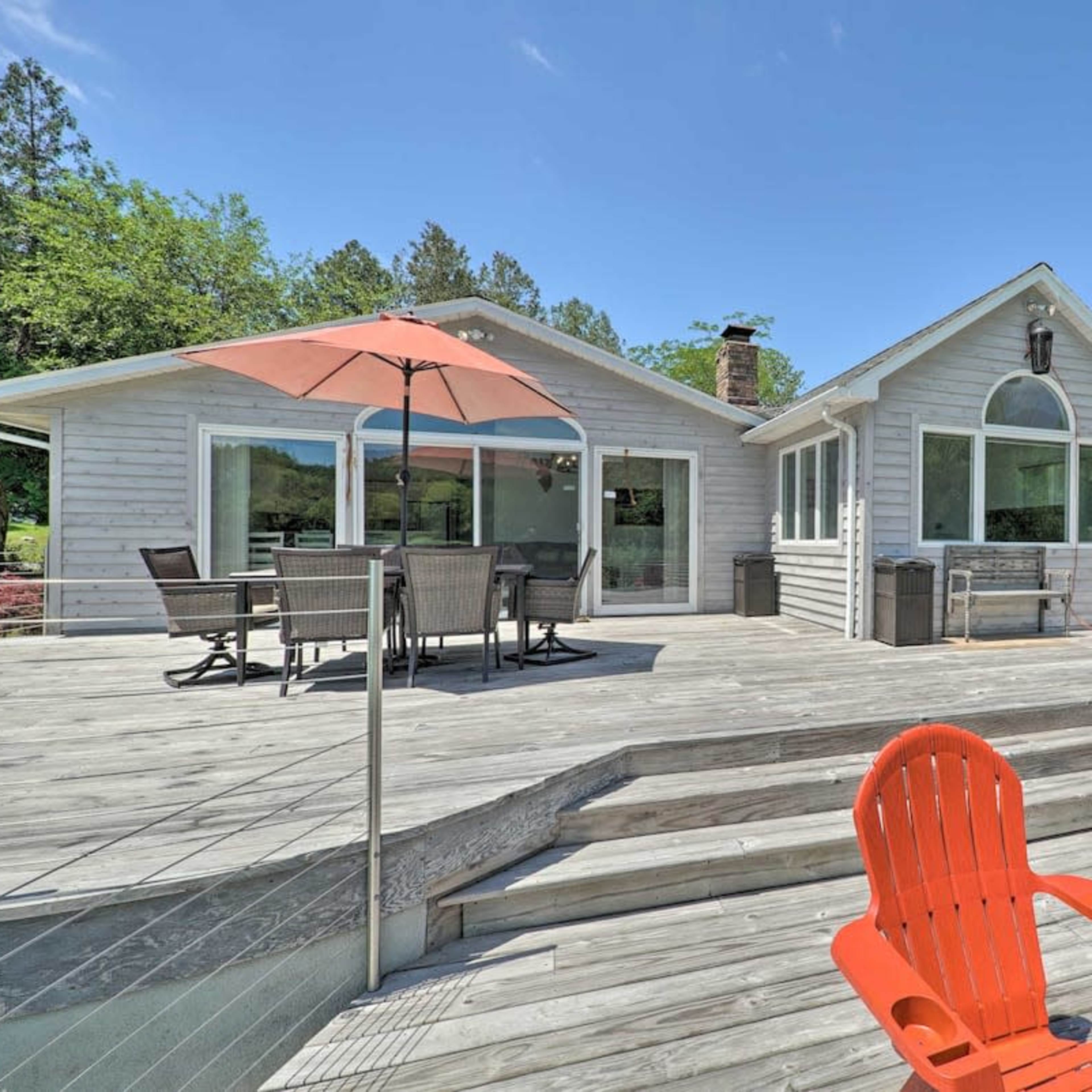 A large wooden deck with two orange chairs and a dining area, adjacent to a modern house surrounded by trees and clear blue skies.