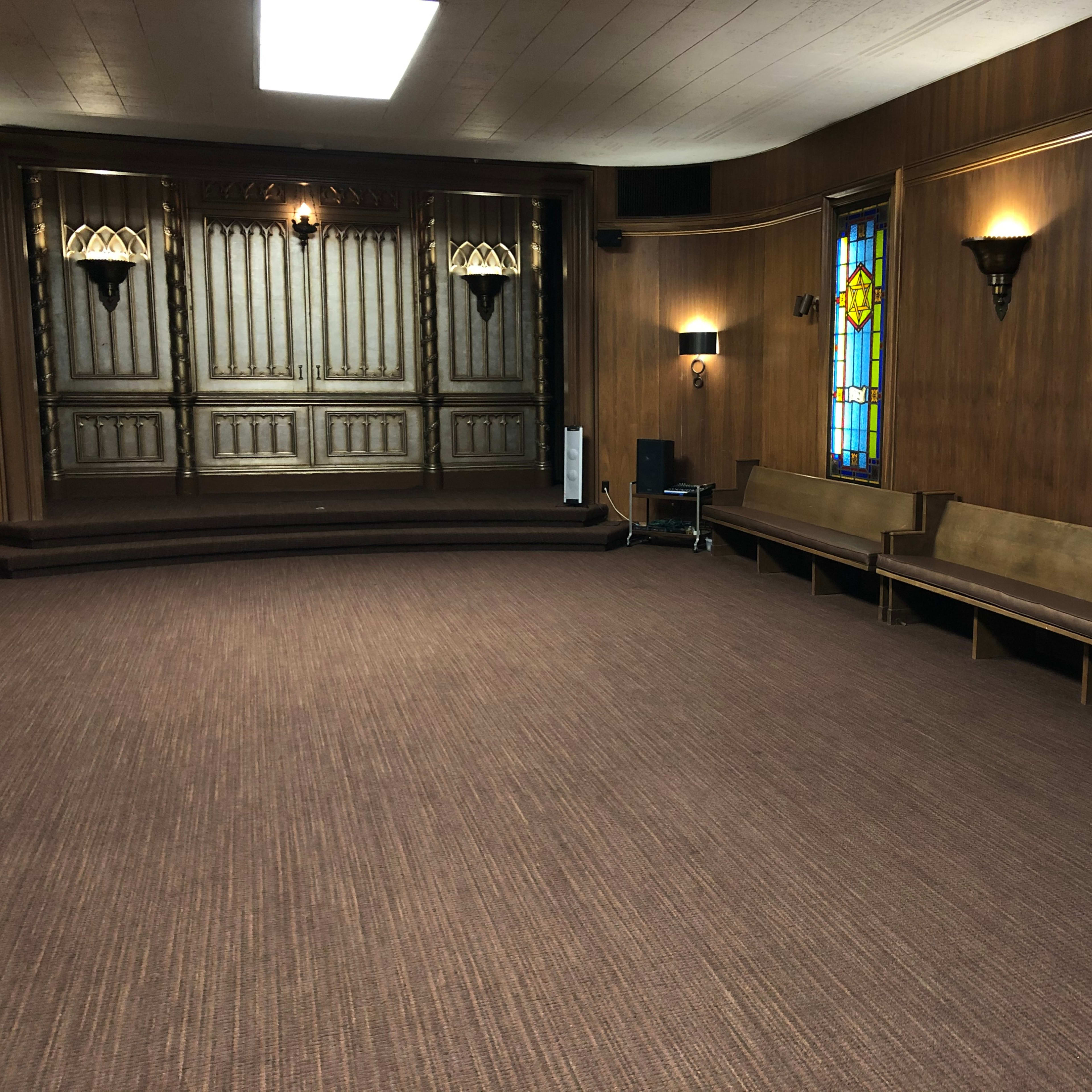 An interior space with wooden walls, benches along the sides, and stained glass windows, featuring a raised stage at the back.