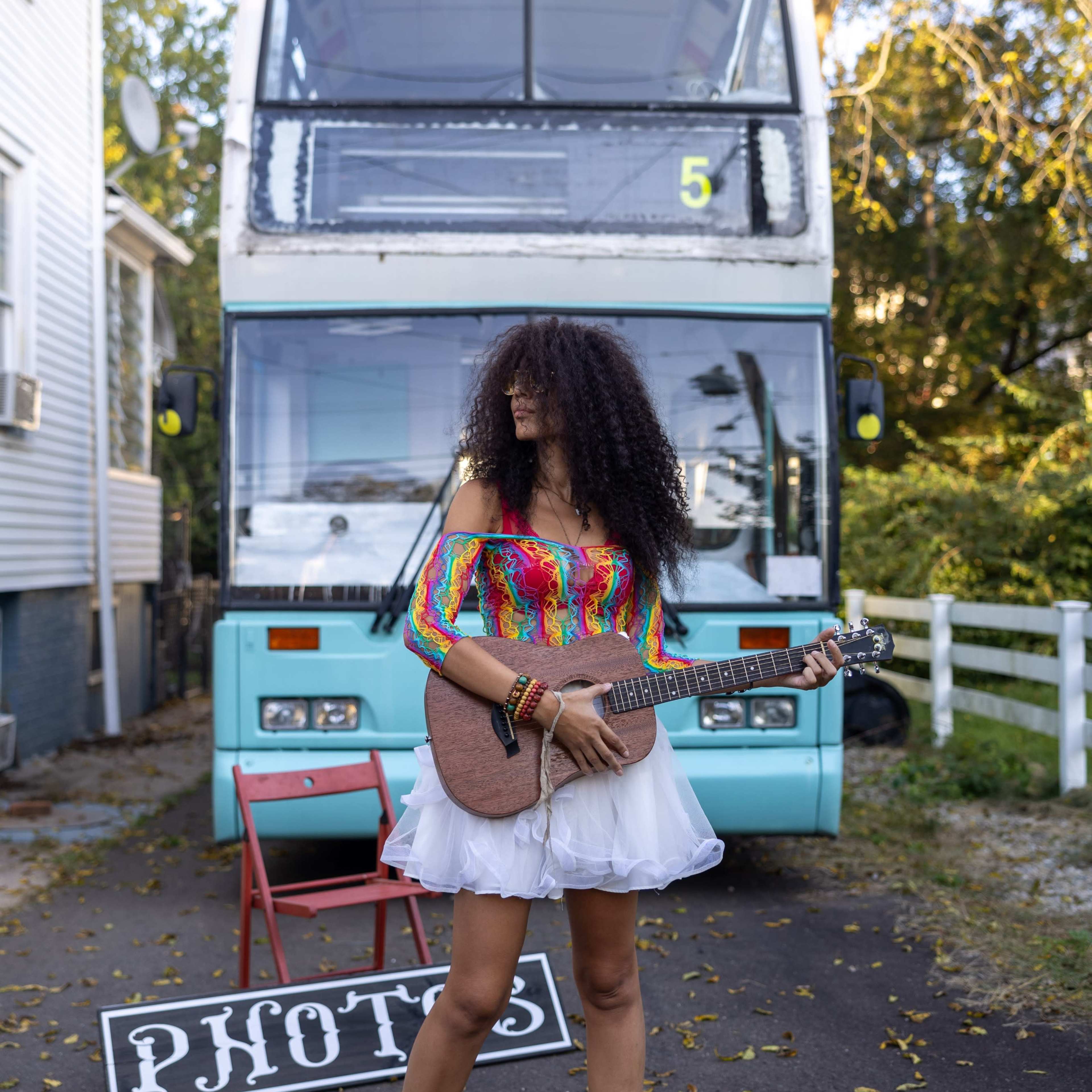 A woman plays a guitar in front of a turquoise double-decker bus, wearing a colorful top and a white skirt.