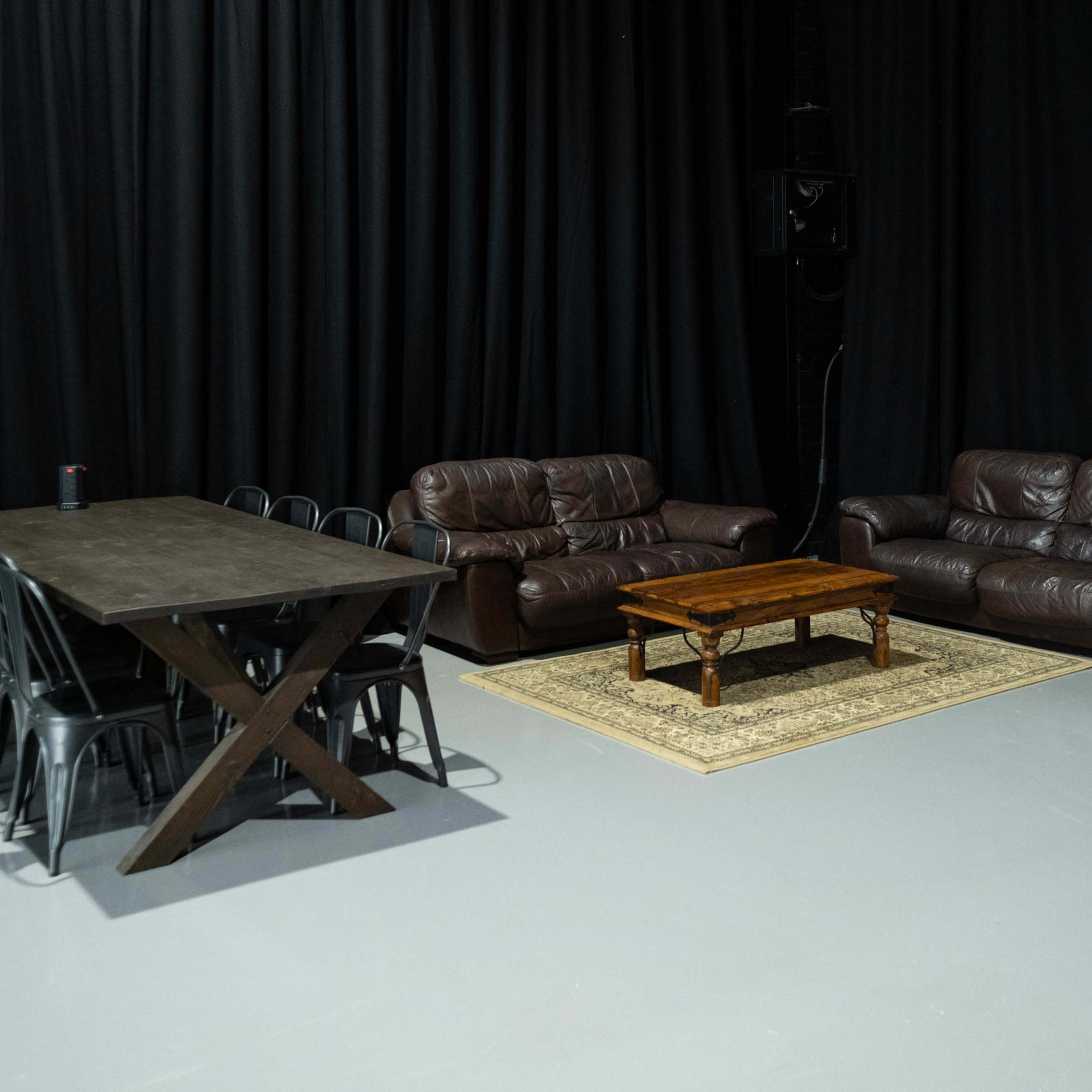 A black-curtained room features a wooden dining table with metal chairs on one side and two leather sofas with a coffee table and a rug on the other.