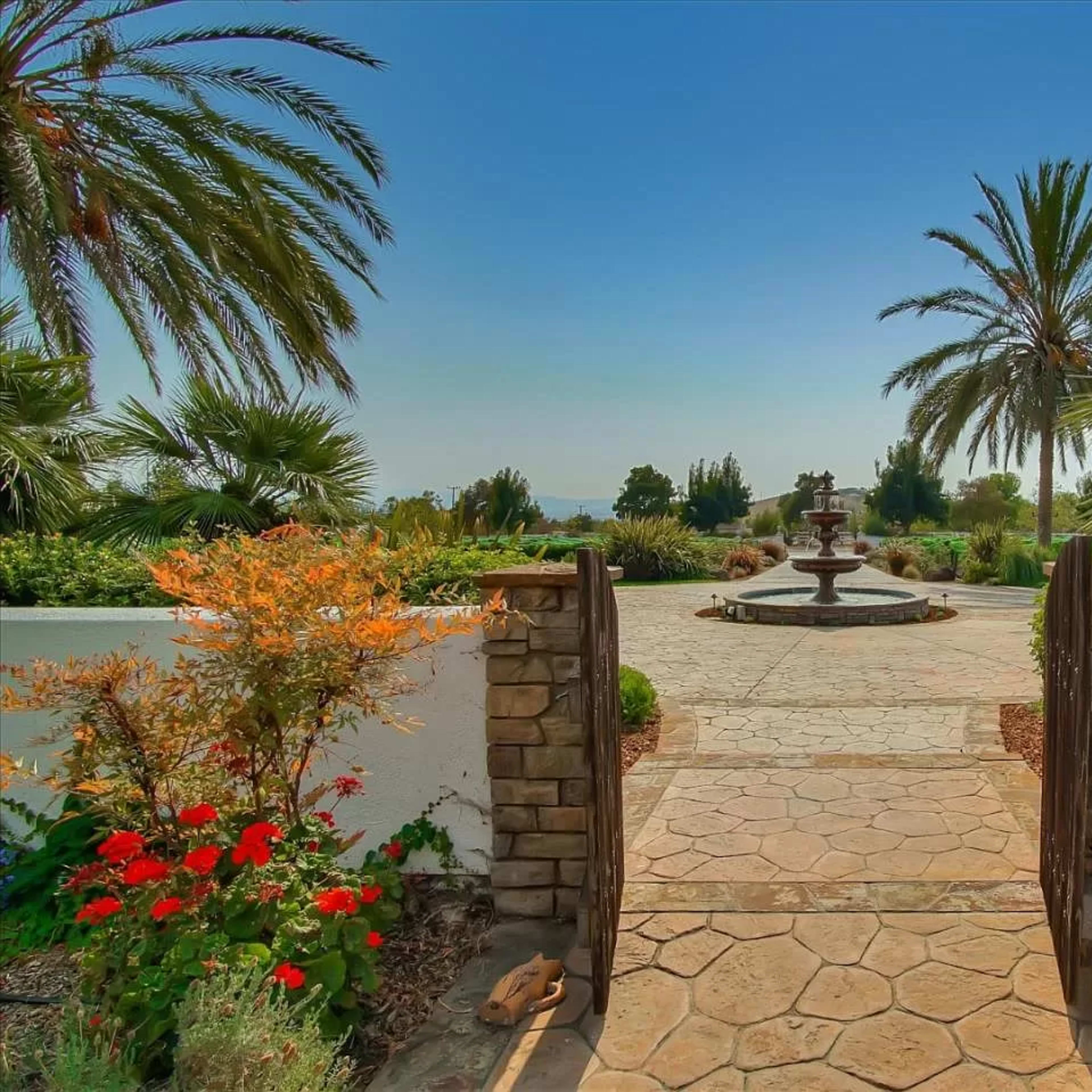 A stone pathway leads through a garden gate to a landscaped area featuring a fountain and palm trees.
