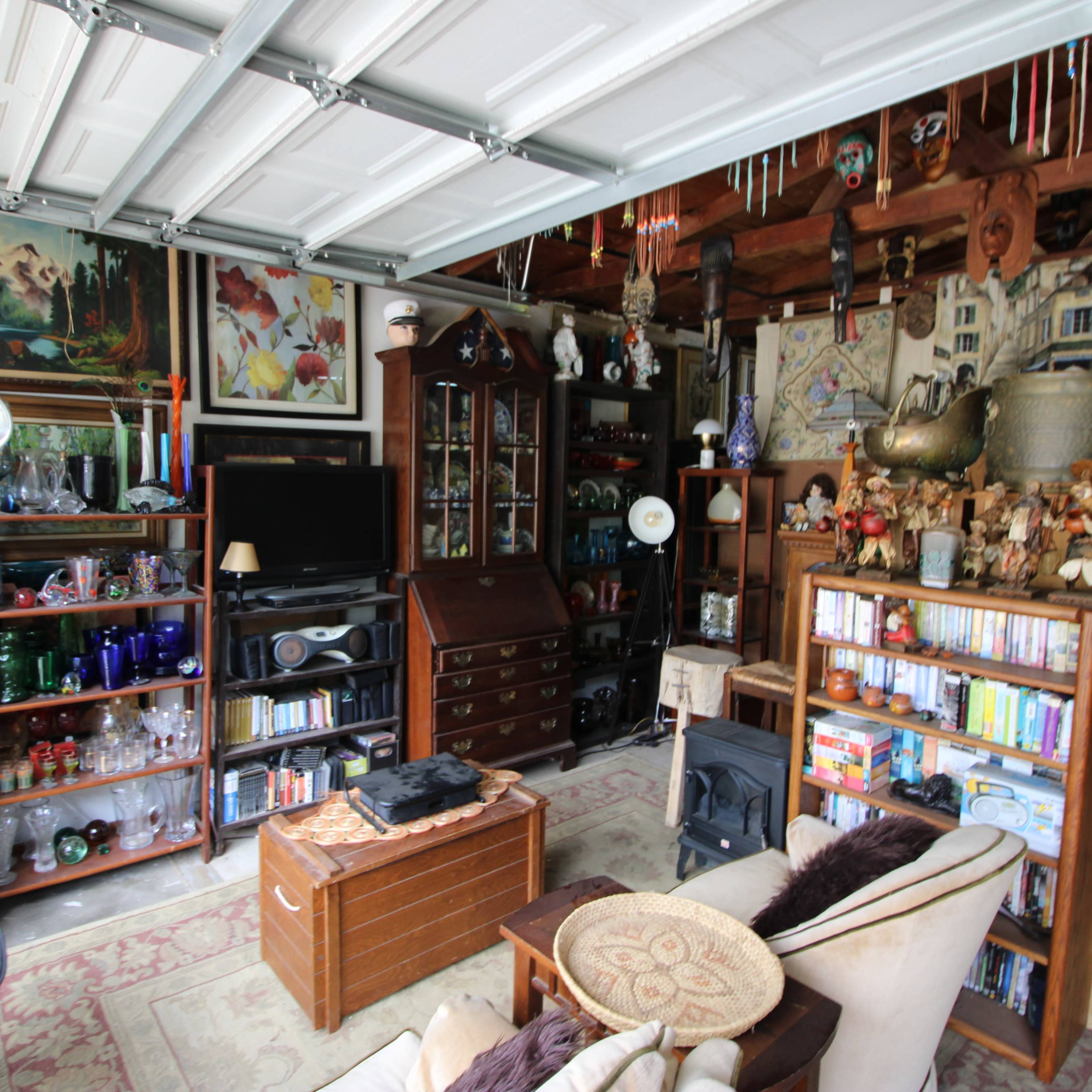 A cluttered garage filled with shelves of books, decorative items, and various artworks, including paintings and sculptures.